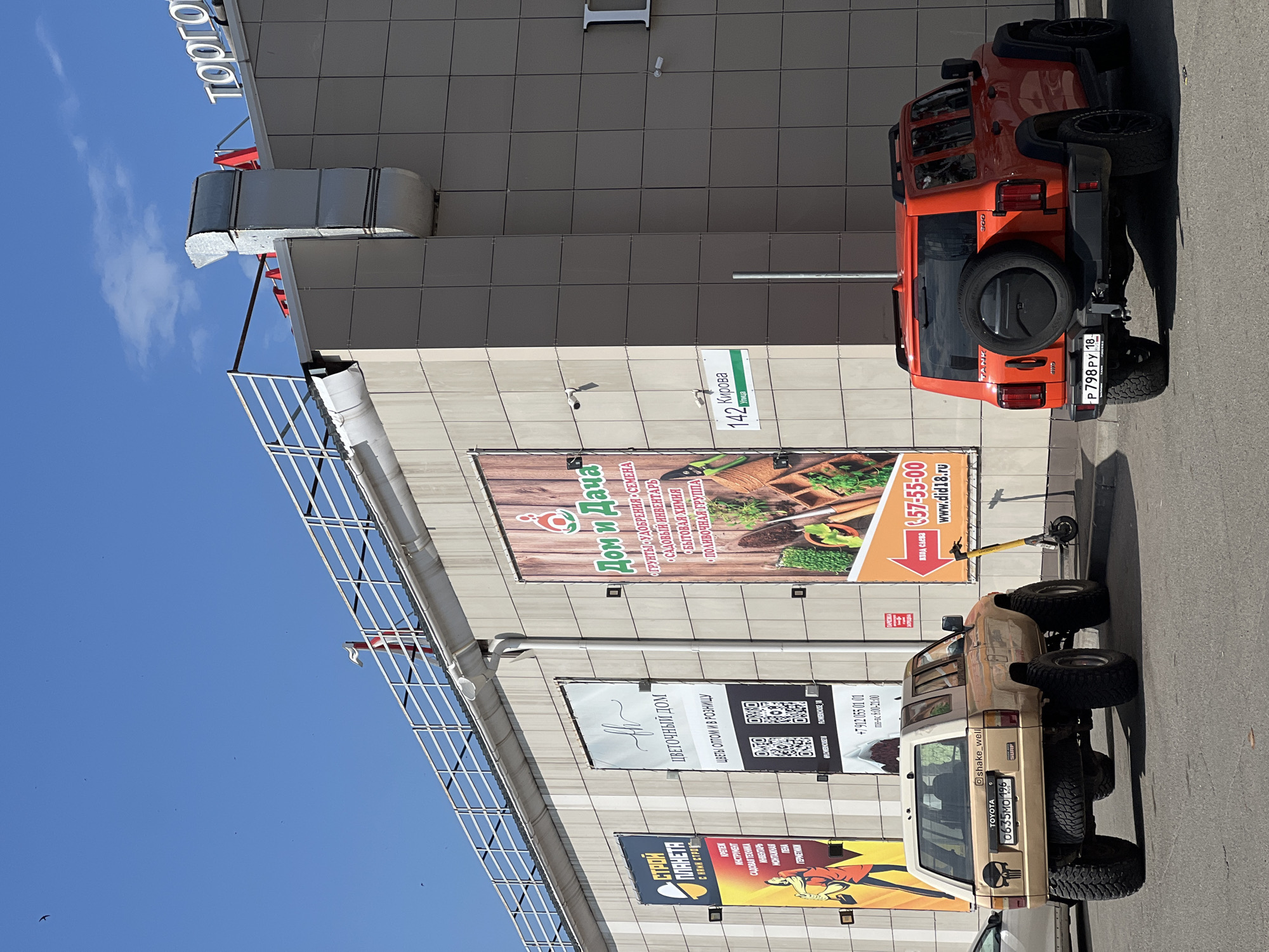 The image shows a bright, sunny day in front of a modern shopping center in Izhevsk, Russia. The shopping center has a facade of beige tiles and large advertising banners. One banner is for "Stroy Planeta" (Construction Planet), another for "Tsvetochiy Dom" (Flower House), and a prominent one for "Dom i Dacha" (House and Dacha), advertising gardening supplies. A street sign for "142 Kirova Ulitsa" is visible.
Two SUVs are parked in front of the shopping center. The vehicle on the left is a tan Toyota 4Runner with large off-road tires and a skull decal on the rear. The vehicle on the right is an orange Tank 300 SUV. A yellow electric scooter is parked next to the "Dom i Dacha" banner. The sky is clear blue with a few small birds visible.