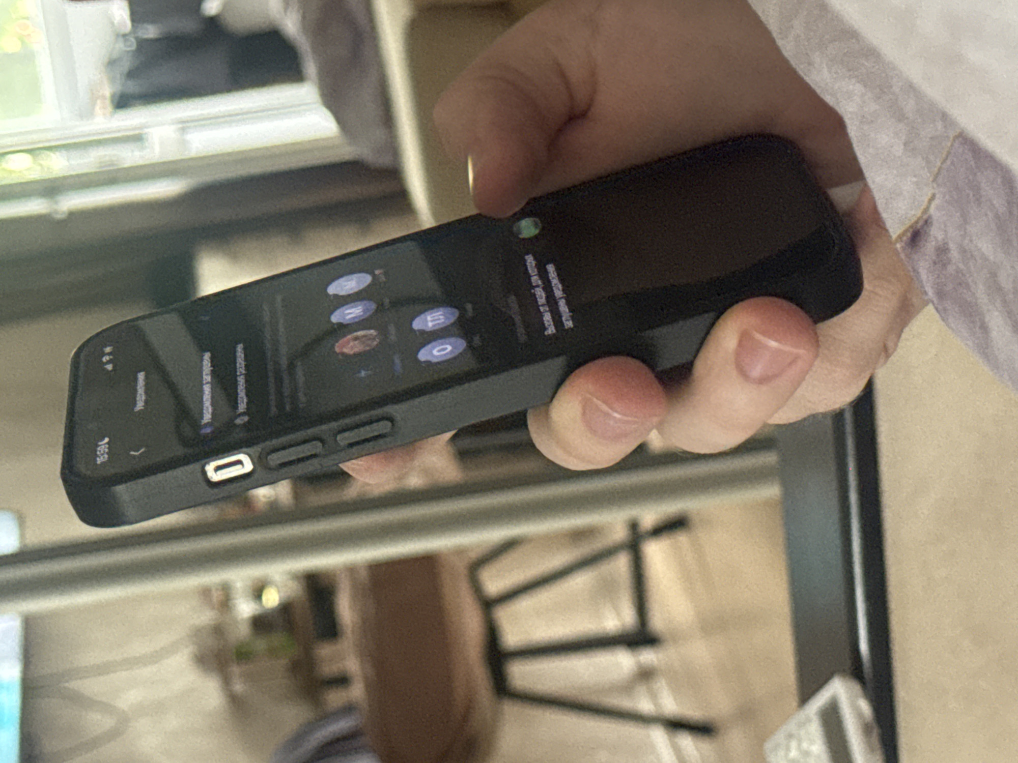 The image shows a close-up of a person holding a black smartphone horizontally. The phone's screen displays a list of notifications and a green indicator light at the top. The background is blurred but suggests an indoor setting with furniture and possibly a window with natural light. The person's hands are visible, with fingers interacting with the phone. Text on the screen is in Russian, mentioning "notifications" and "calls from people for whom notifications are muted." The time displayed on the phone is 16:59. The overall scene appears casual, likely a person checking their phone in a home or office environment.