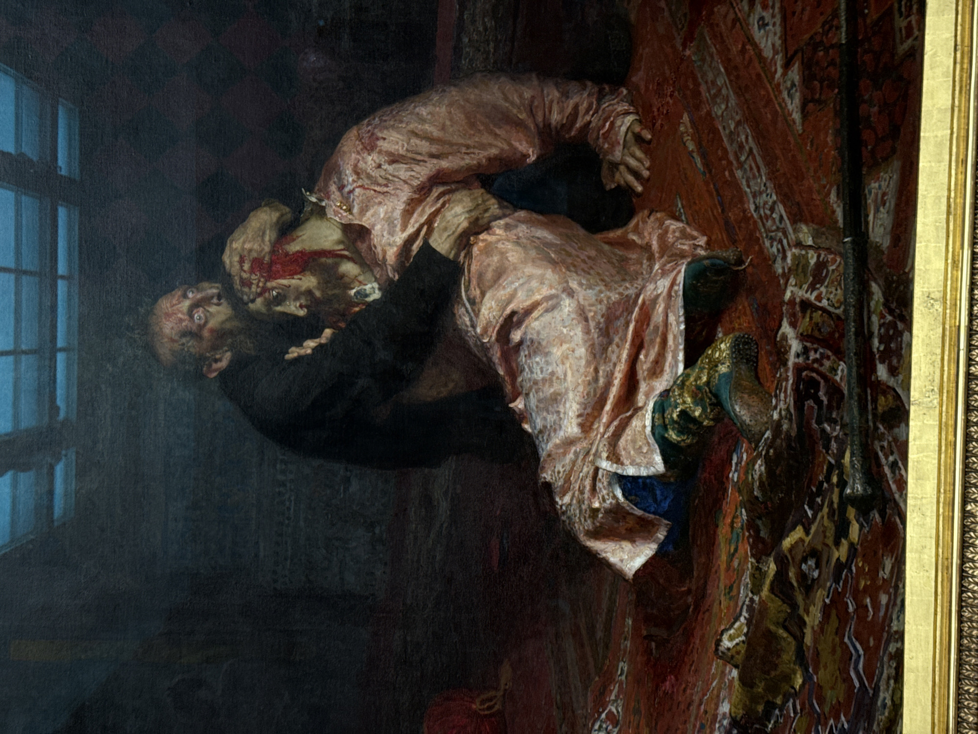 The image is a painting depicting a murder scene in Moscow, Russia. Two men are visible. One man, dressed in a dark suit, is strangling another man wearing a pink robe. Both men appear to be bleeding from head wounds. The scene takes place in a room with a window on the left and a patterned rug on the floor. A rifle is leaning against the wall next to the rug. The scene is dark and violent.