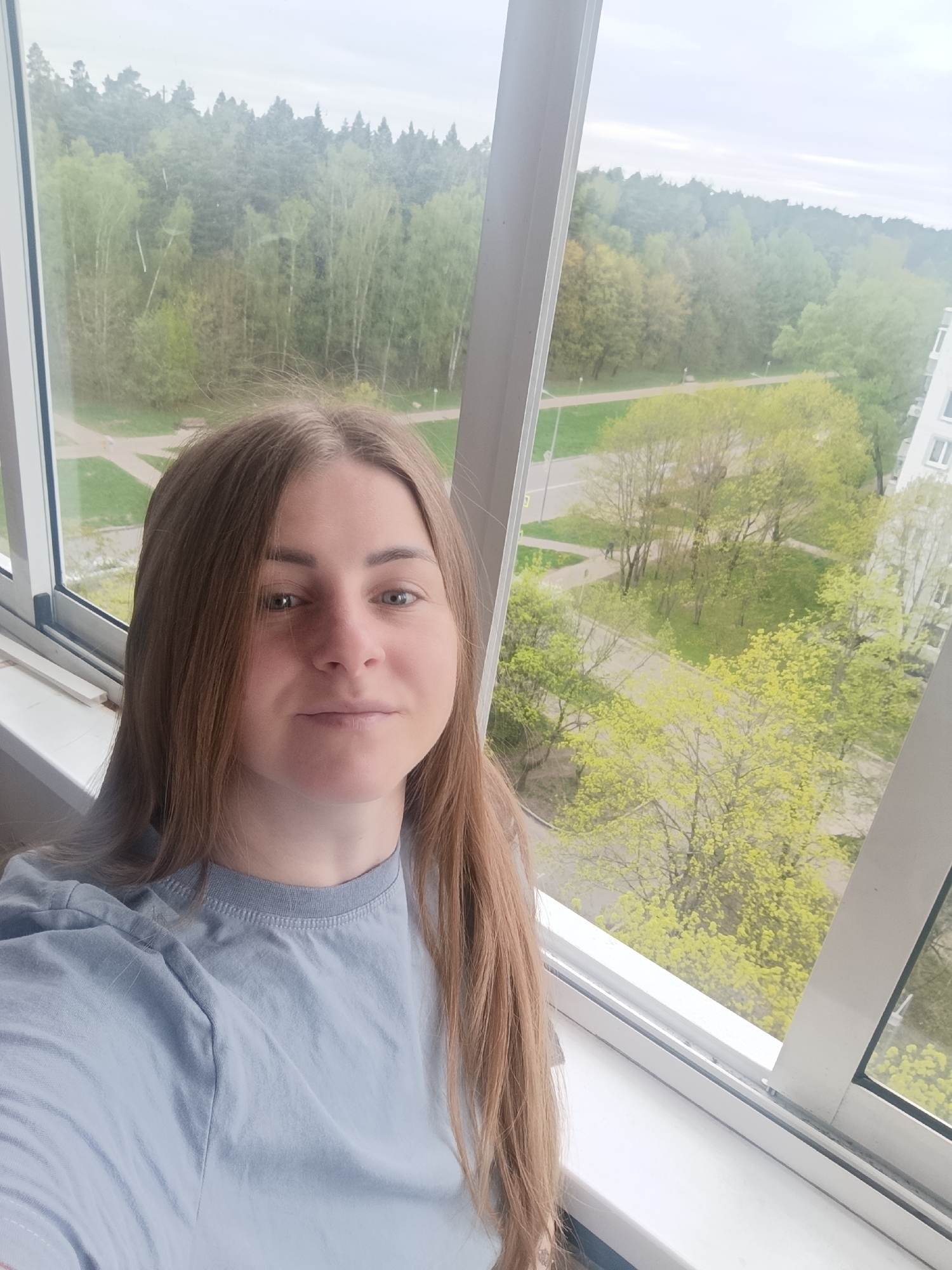 A medium-angle shot shows a woman with long, straight, light-brown hair in front of a large window. She is wearing a light blue T-shirt. Outside the window is a view of trees with green leaves, sidewalks, buildings, and other trees in the distance. The weather is overcast.