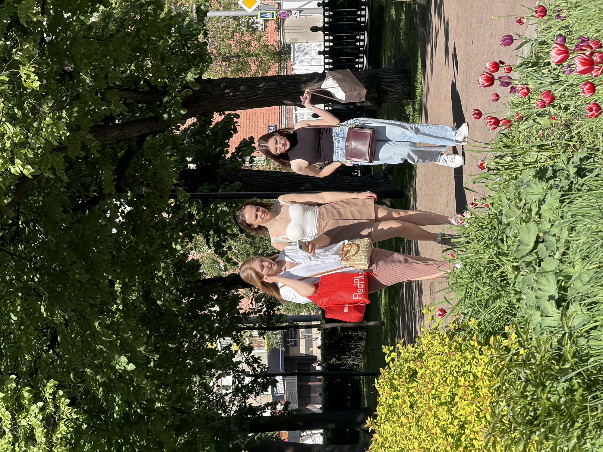 Three young women are walking on a sidewalk in a park. The woman nearest the camera is wearing a white shirt, pink skirt, and has a red bag labeled "RedPlus". The woman in the middle wears a white top and brown skirt, while the woman farthest from the camera is wearing a black top and jeans and carrying a shopping bag. The women appear to be walking in Moscow, Russia, evidenced by the street signs and building style in the background. Tulips grow on the edge of the sidewalk.