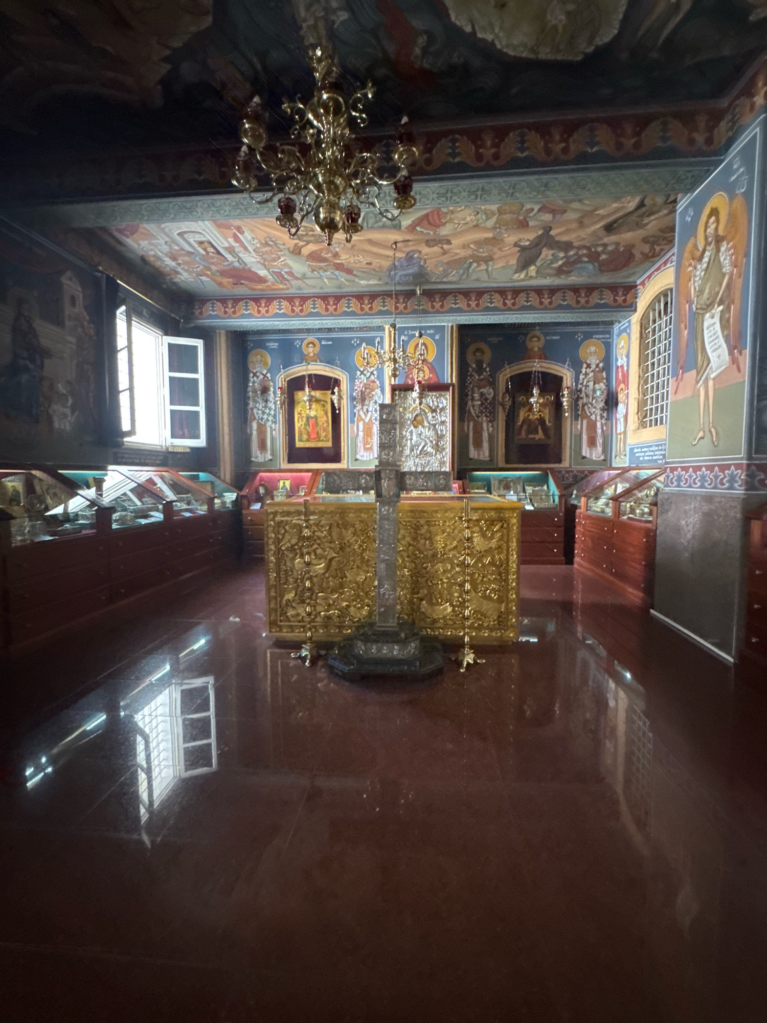 The image shows the interior of a church, likely the Church of Ayios Ioannis Prodromos in Tsakistra, Cyprus. The scene is dominated by religious iconography and opulent decor. In the center, a large, ornate golden structure, possibly an altar or iconostasis, is adorned with intricate carvings and stands before a backdrop of religious paintings and icons on the blue walls. A tall, metallic cross is positioned in front of the golden structure.
The ceiling is covered with religious murals, depicting various biblical scenes, and a decorative chandelier hangs down. To the left and right of the central altar area, glass display cases filled with religious artifacts are visible, suggesting this area might be a museum or exhibition space within the church. Natural light enters through a window on the left side of the image, casting reflections on the polished, dark red floor. The overall impression is one of solemnity and rich spiritual heritage.