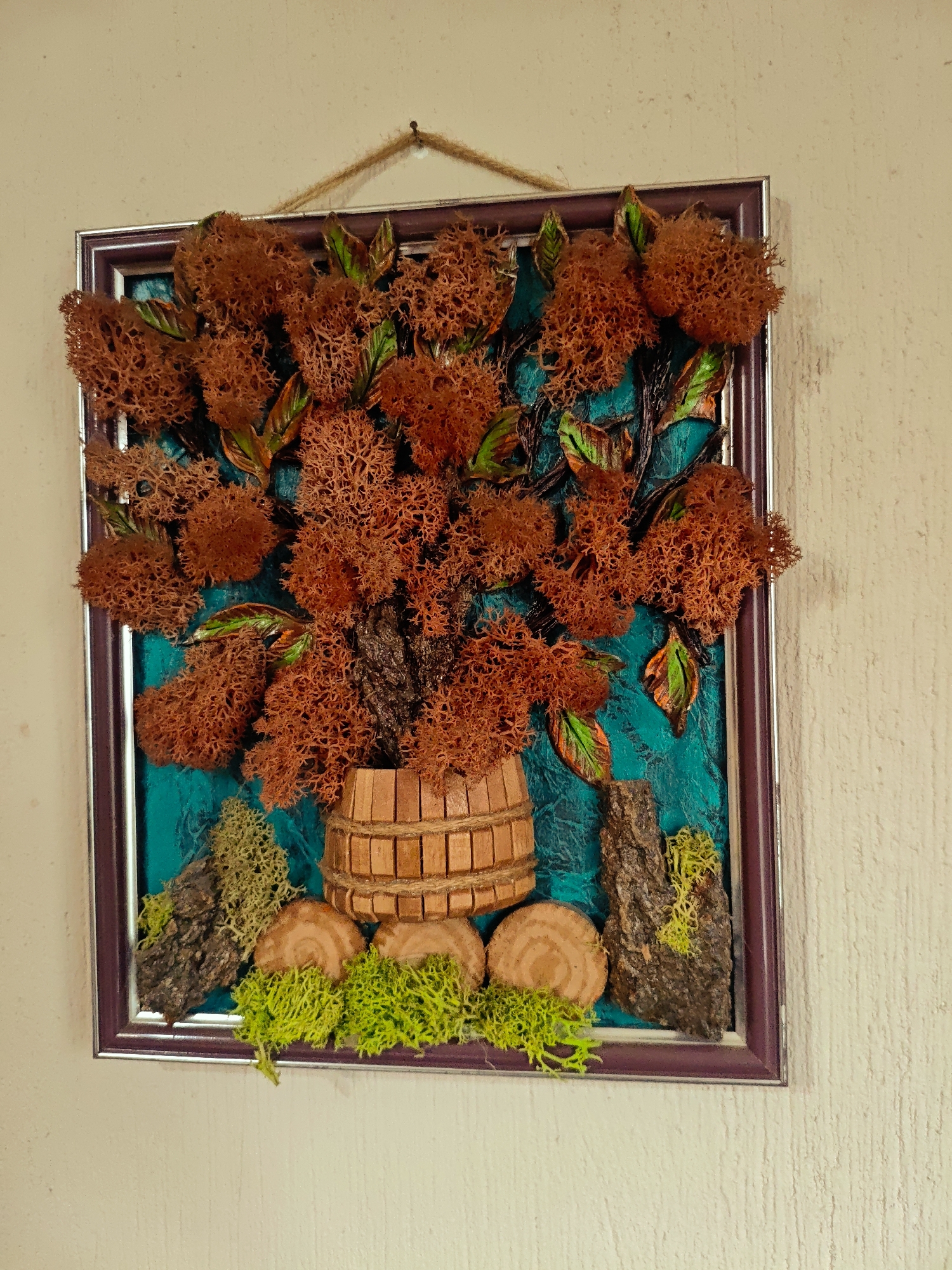 A framed wall-mounted floral arrangement hangs against a light beige, textured wall. The arrangement features a rustic, wooden barrel-shaped container filled with a brown, moss-like material forming a bush or tree. Green leaves are interspersed among the brown moss. The background is a painted, teal-colored surface. At the base of the barrel are wooden discs and tufts of bright green moss, along with pieces of bark. The frame is two-toned, with a silver outer edge and a dark purple inner edge. The frame is suspended by a simple brown string or cord. The location is unspecified.