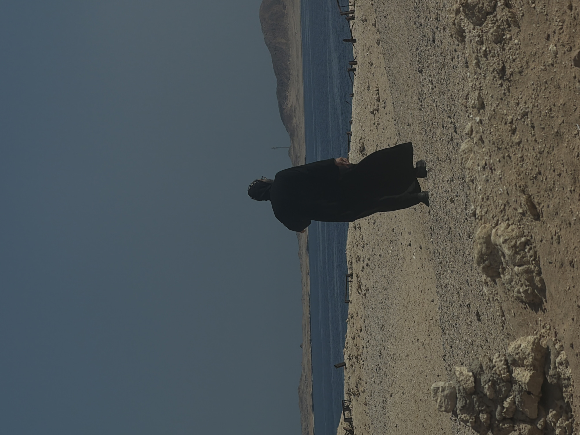 A person wearing a black abaya and headscarf walks across a sandy, rocky shore towards the sea under a clear blue sky. The sea is calm and blue, with a distant sandy coastline and mountains. In the foreground, there are small piles of stones. The scene appears to be in a coastal area of Sharm El Sheikh, Egypt.