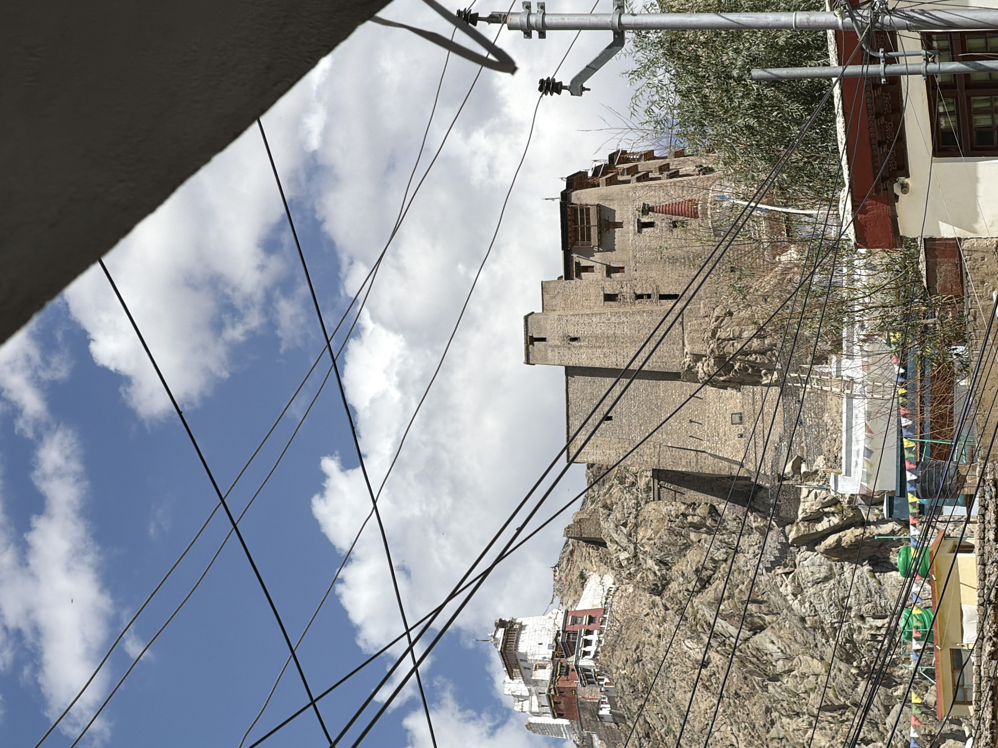 The image depicts the Leh Palace, a historic 17th-century royal palace located in Leh, Ladakh, India. The palace is situated on a rocky hill, with the vast mountainous terrain of the Himalayas serving as the backdrop. The sky above is a vibrant blue, dotted with fluffy white clouds.
In the foreground, a tangled network of electrical wires and utility poles dominates the view, partially obscuring the palace and the surrounding landscape. These wires create a modern, urban overlay on the otherwise ancient and natural scene.
The Leh Palace itself is a multi-story structure, characterized by its imposing stone walls and traditional Tibetan architecture. Sections of the palace are painted white, contrasting with the earthy tones of the rock face. Smaller buildings, possibly residential or administrative, are visible at the base of the hill. Prayer flags, a common sight in Buddhist regions, are strung across some of the lower structures, adding a splash of color.
To the right of the palace, the frame is partially filled by a modern building with a white facade and a red-trimmed roof. A prominent utility pole with insulators and multiple wires stands adjacent to this building, further emphasizing the juxtaposition of the old and the new in the urban environment of Leh. The overall scene captures the unique blend of history, culture, and modern infrastructure found in this high-altitude city.