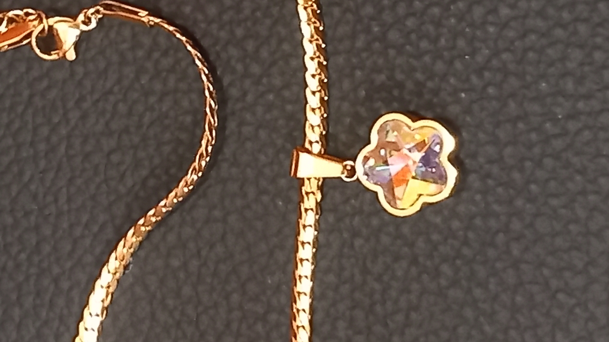 A close-up, eye-level shot shows a gold chain with a small, flower-shaped pendant. The pendant contains a faceted, multi-colored gemstone. The background is a dark, textured surface, likely leather or a similar material. The image is well-lit, highlighting the shine of the gold and the sparkle of the gemstone. The scene appears to be a product photograph or a personal item display. No specific location in the city of Kos, Greece, can be identified from the image.