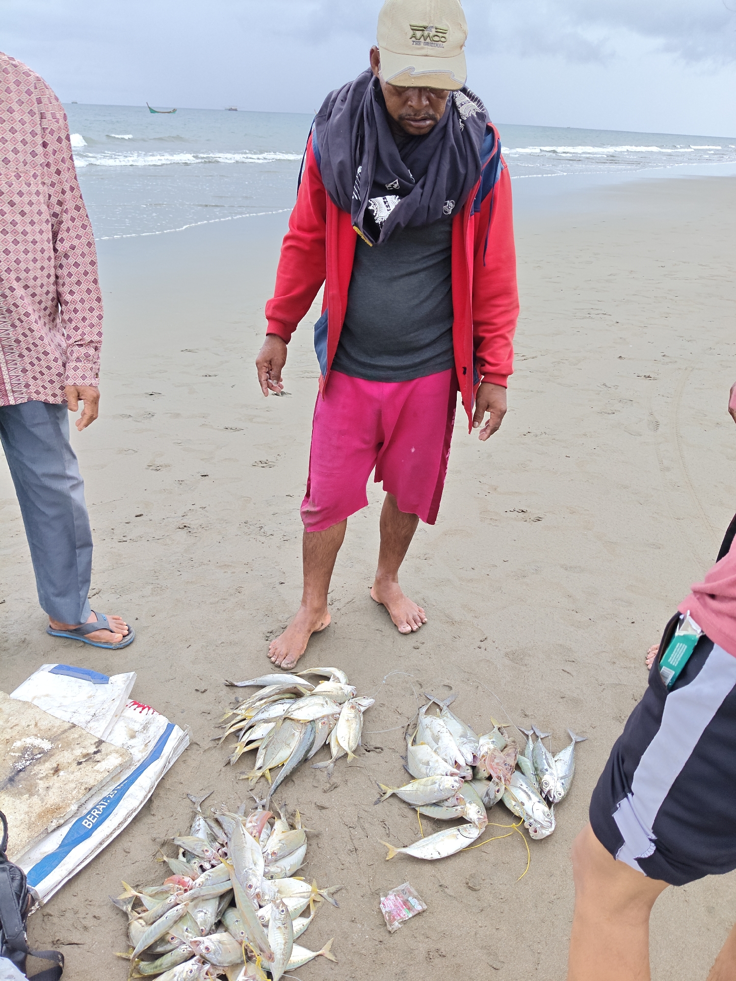 In Curehtunong, Indonesia, on a sandy beach with the ocean in the background, three individuals are present. The person in the center, wearing a red jacket, gray t-shirt, and pink shorts, stands over two piles of small fish laid out on the sand. To the left, another individual, wearing a patterned shirt and gray pants, stands with their feet in flip-flops. A third person, only their lower legs and feet visible, is on the right, wearing black and white shorts. One pile of fish is closer to the foreground, while another is slightly further back. A white and blue tarp-like object is situated to the left of the fish. The sky is overcast.