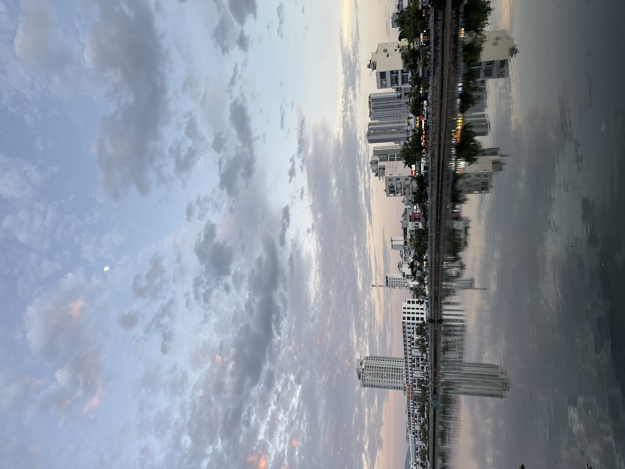 The image captures a serene view of the Nha Trang cityscape at dusk, with a prominent river reflecting the illuminated buildings and the soft hues of the sky. The sky is adorned with a scattering of clouds, tinged with pink and gray, and a pale moon is visible. Along the riverbank, modern high-rise buildings and lower-rise structures form a cityscape, their lights beginning to twinkle. Trees line the riverfront, adding a touch of greenery. The river itself is calm, its surface acting as a mirror, doubling the visual impression of the buildings and the sky. The overall atmosphere is tranquil, suggesting the transition from day to night in this Vietnamese coastal city.