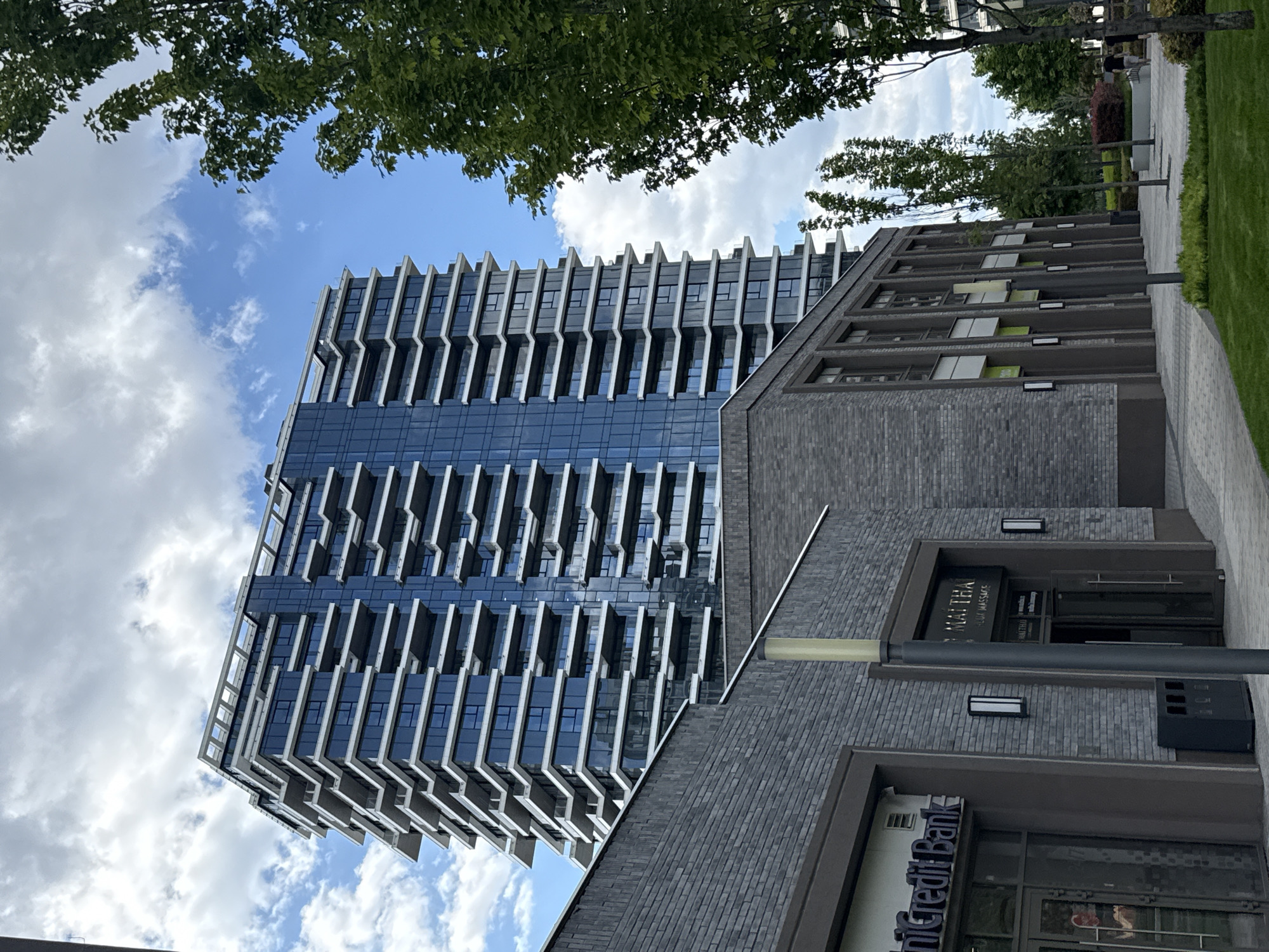 The image depicts a modern architectural scene in Belgrade, Serbia, featuring a tall, glass-clad residential or commercial tower with repeating cantilevered balconies. The building's design emphasizes verticality, with the balconies creating a rhythmic, stepped appearance. To the right of the tower, a lower, brick-clad structure is visible, housing street-level businesses. Based on signage, these include a UniCredit Bank and what appears to be a massage or spa establishment named "Kaltra". The view is from a pedestrian perspective, possibly on a sidewalk. The upper portion of the frame is partially obscured by tree foliage, and a blue sky with scattered clouds provides the backdrop. No individuals are clearly visible in the scene.