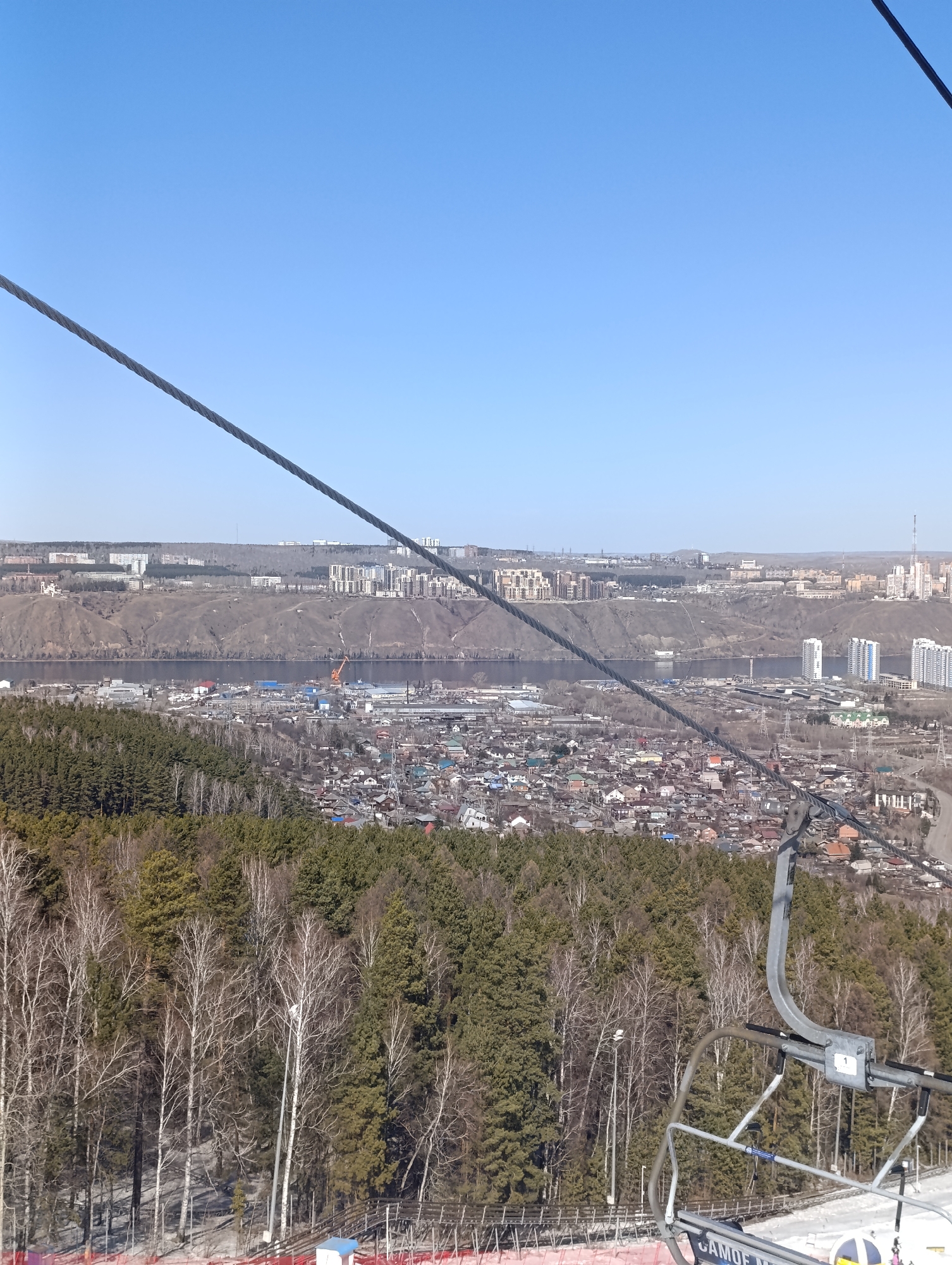 This eye-level shot overlooks the cityscape of Krasnoyarsk, Russia, from a high vantage point, likely a ski lift. The foreground is filled with a dense forest, primarily composed of coniferous and deciduous trees. Traces of snow are visible on the ground, indicating a late winter or early spring setting. An industrial area is visible along the river, and residential areas stretch across the landscape. The background shows the distant skyline of Krasnoyarsk, with high-rise buildings and structures. The sky is a clear, bright blue. The ski lift's cable and part of its chair are prominently featured, adding depth to the composition and confirming the elevated perspective.