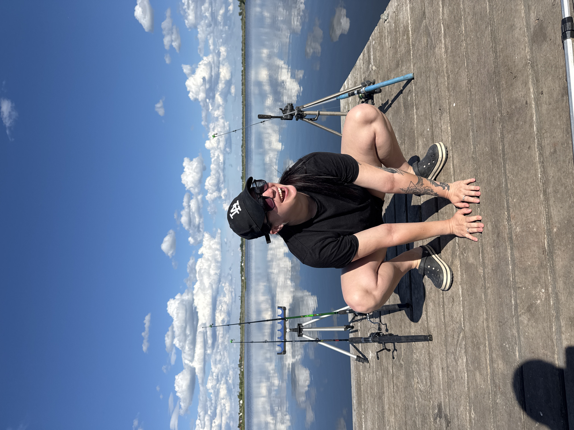 On a sunny day in Unknown, Unknown, a woman wearing a black t-shirt, sunglasses, and a black baseball cap squats on a wooden dock. Two fishing rods stand behind her on tripods. The sky above is blue with fluffy white clouds that are reflected in the water behind the dock. The woman is smiling and looks to the left. She wears dark colored Crocs-style shoes and has tattoos on her arms.