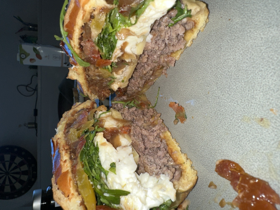 A large burger, sliced cleanly through its horizontal axis, rests on a light gray, textured plate. The burger's interior is exposed, revealing a thick, medium-cooked ground beef patty with a pinkish-brown center. Above the patty, a substantial layer of white, creamy cheese (consistent with mozzarella or burrata) is visible, topped with a generous quantity of green leafy vegetables, likely arugula. Further up, layers of caramelized onions, sliced yellow peppers or pickles, and a reddish sauce are present between the toasted bun halves. Minor drips and smears of sauce, containing diced red elements, are evident on the plate surrounding the burger. The background is dimly lit and out of focus, revealing an indoor setting. On the left, a dark dartboard with concentric circles and radial divisions hangs on a wall. To the right of the dartboard, a dark-lidded jar and a white carton container with graphics are discernible on a surface. The scene is located in Carnin, France.
