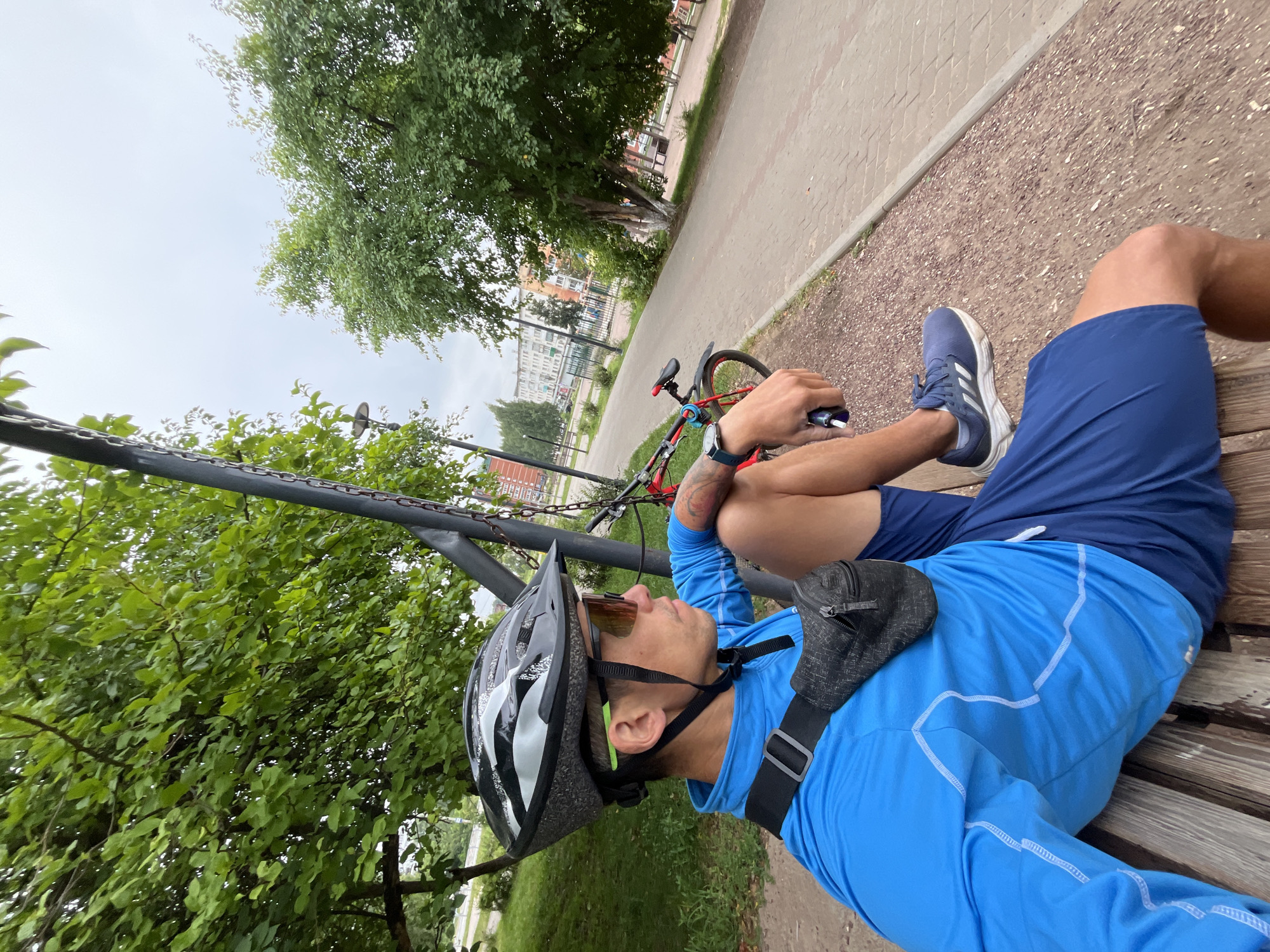 A man wearing a blue long-sleeved athletic shirt, dark blue shorts, a black cycling helmet, and orange-tinted sunglasses sits on a wooden park bench in Chaikovsky, Russia. He has a dark gray fanny pack around his waist. To his right, a red bicycle is parked upright. The man is looking to his right. The background shows a paved walkway, green trees, and apartment buildings. The sky is overcast.