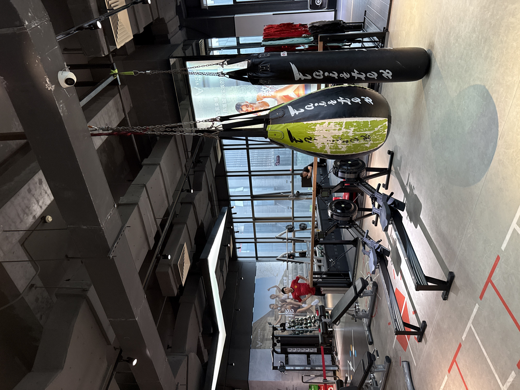 This is a medium shot of the interior of a modern gym, likely located in Haizhu District, China, as evidenced by the signage visible in the background. The gym features an industrial aesthetic with exposed ductwork and concrete ceilings. In the foreground, two Concept2 rowing machines are positioned on a light gray floor marked with red and white lines. To the right and slightly behind the rowing machines, two punching bags hang from the ceiling by chains; one is black and the other is a split design of black and distressed lime green. In the mid-ground, a man in a red shirt and black shorts stands facing the camera, posing in front of a large graphic display featuring images of athletes. Behind him, various weight training equipment, including a squat rack with weights and a multi-functional exercise machine, are visible. To the far right, a rack of red and white towels hangs on the wall. The background reveals large windows offering a view of a city skyline with tall buildings. The lighting is a mix of ambient light from the windows and artificial track lighting on the ceiling.
