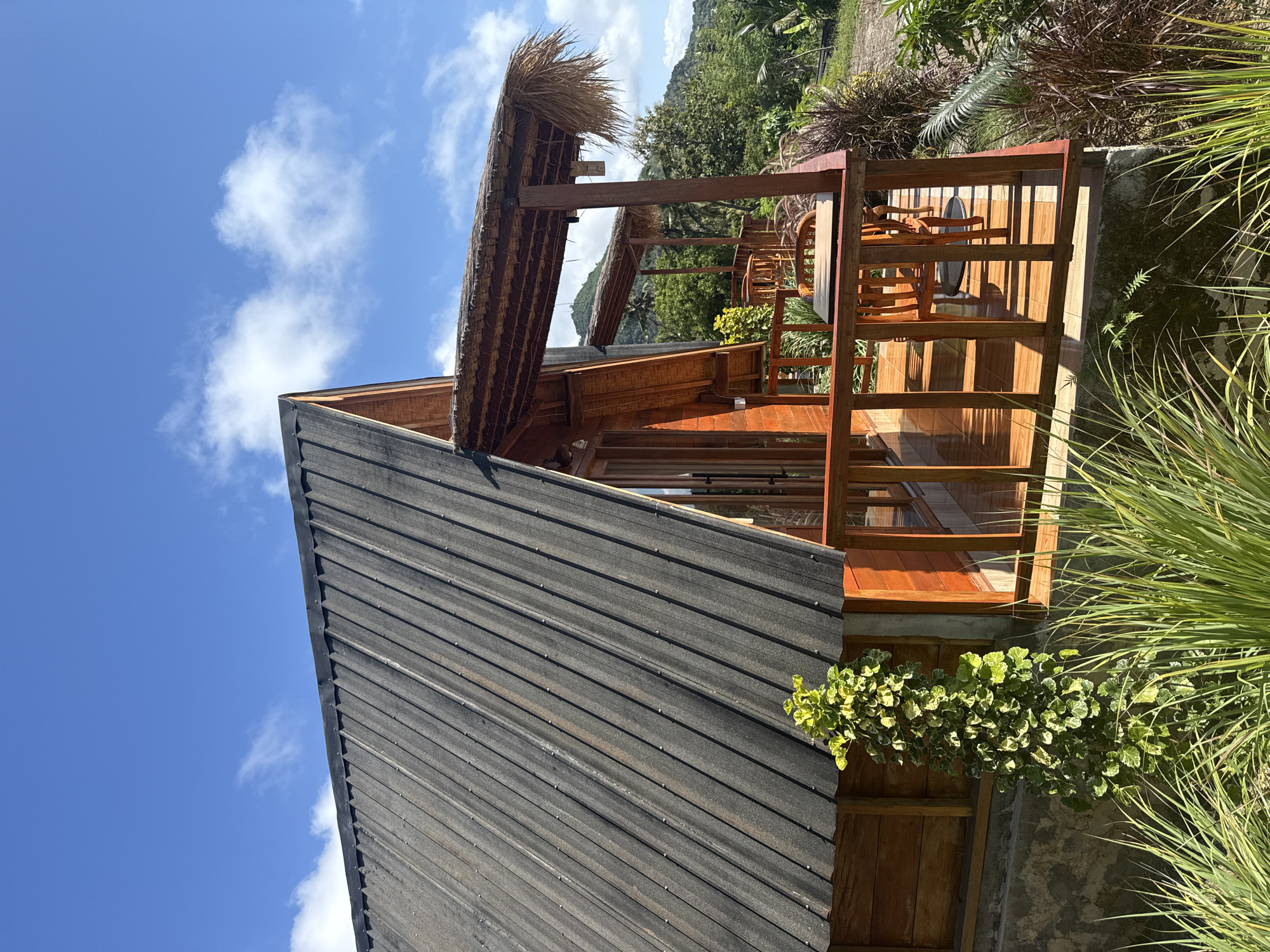 Exterior shot of a wooden cabin located in Tanglad, Indonesia. The cabin has a black metal roof, wooden walls and a thatched roof extension with a wooden deck. The sky is blue with some clouds. Greenery is around the cabin.