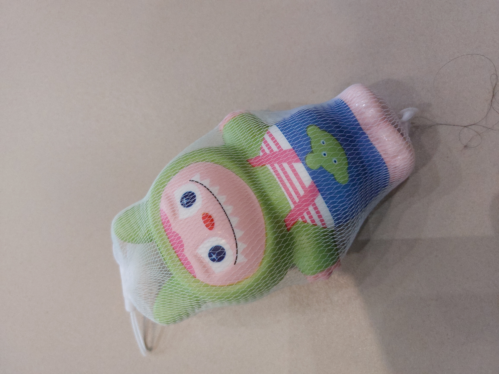 A toy character, which is green with pink accents, is encased in a white mesh bag. The toy has large blue eyes, a wide mouth with sharp teeth, and is wearing blue overalls with pink straps. A small green alien-like figure is visible on the overalls. The toy is lying on a light-brown, textured surface. A few strands of dark hair are on the surface near the toy. The image is a close-up shot of the toy. The location in the city of Подкумок, Russia, is not discernible from the provided image.