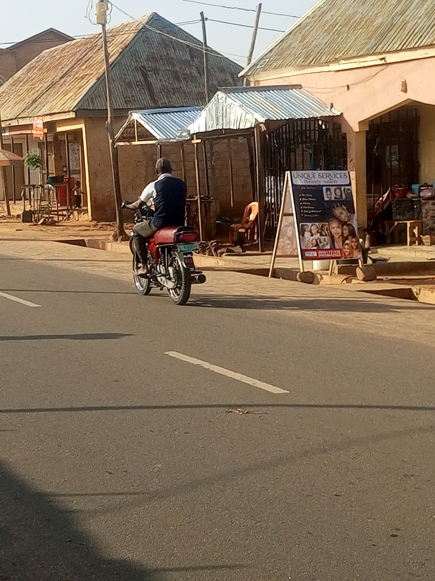 The image shows a street scene in Tempure, Nigeria, on a sunny day. A man is riding a red motorcycle on the right side of the road. He is wearing a white shirt and a dark vest. The motorcycle has a license plate with some writing on it. In the background, there are several buildings with corrugated metal roofs. One building has a sign that says "UNIQUE SERVICES Beauty salon". There is also an orange chair and a white easel with a poster on it. The road is paved and has white lane markings.