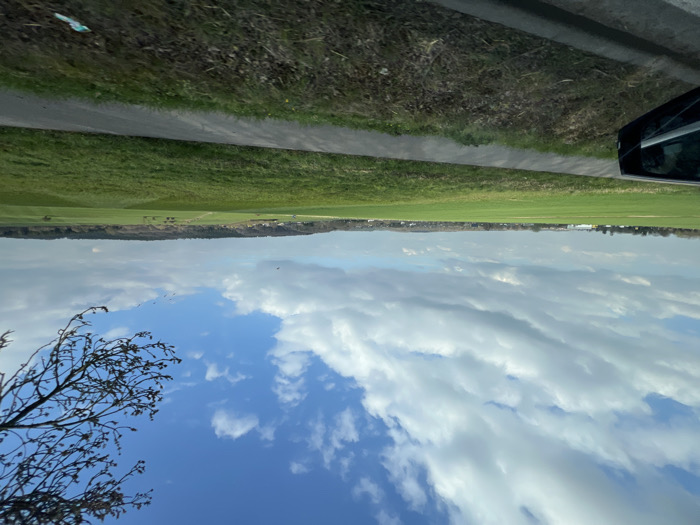 The image displays a panoramic view of an expansive rural landscape within the city of Stolberg, Germany, captured from a low perspective, likely from a moving vehicle. In the immediate foreground, the dark exterior of a vehicle is visible on the left, including a side mirror reflecting a blurred internal scene. A dark grey paved path or road runs horizontally across the lower portion of the frame, bordered by green and brown grassy verges.
The midground and background are dominated by vast, undulating green fields stretching towards a distant horizon. A subtle dirt track or path is observable traversing these fields into the distance. The horizon line reveals a mix of distant structures, including some light-colored buildings with industrial characteristics on the far left, interspersed with dense tree lines suggesting forested areas or hills.
The sky occupies the upper half of the image, featuring a dynamic composition of large, scattered white and grey cumulus clouds against patches of clear blue sky. In the upper right quadrant, bare tree branches with visible small buds extend into the frame. Several small, dark specks, consistent with birds, are observable in the mid-sky. A small piece of light-colored litter is present on the grassy verge in the bottom right corner. No individuals are visible.