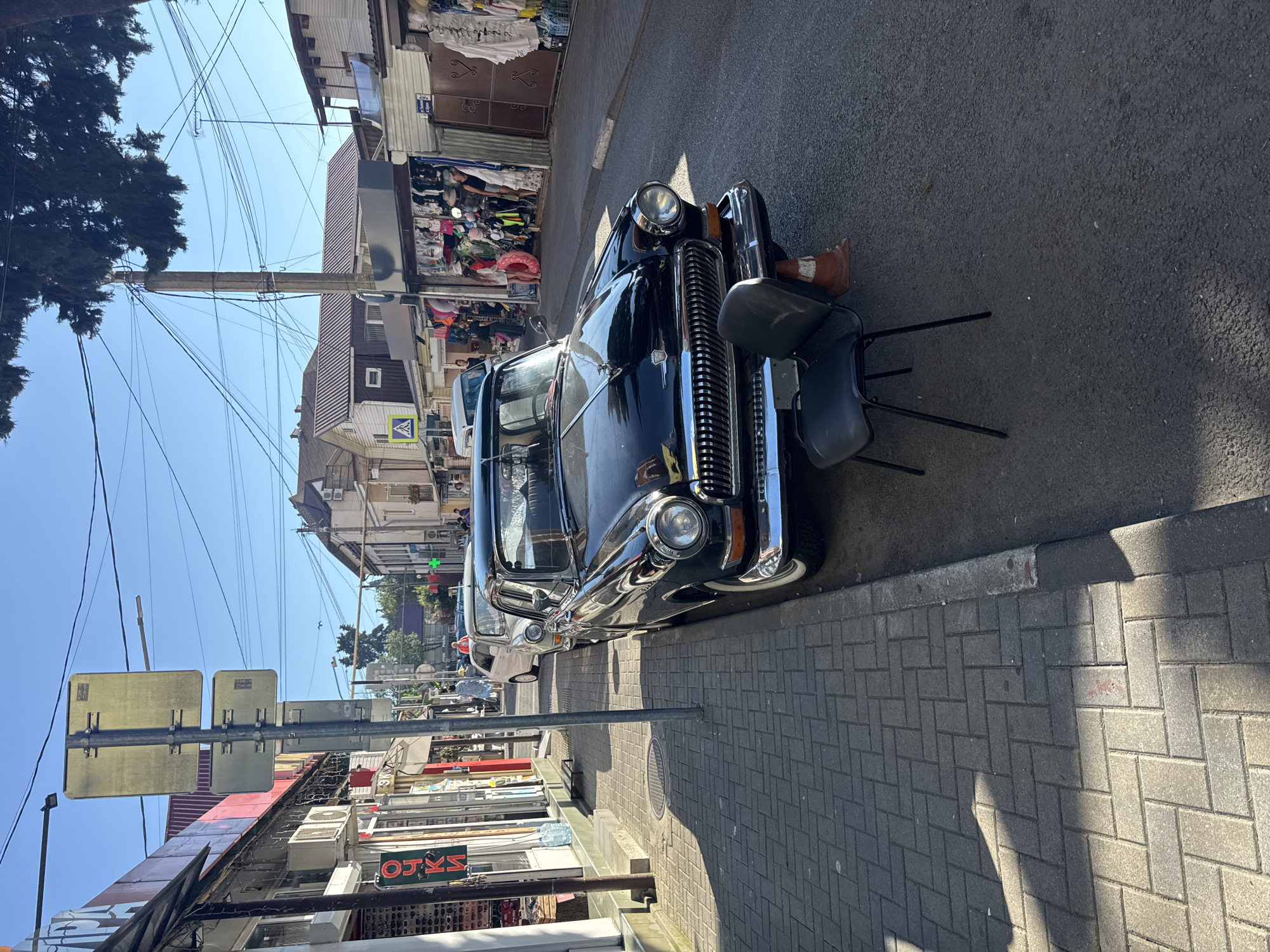The image depicts a street scene in Sochi, Russia, under a clear blue sky. A vintage black car is parked along the side of the road, its chrome grille and headlights gleaming in the sunlight. A black chair and an orange traffic cone are placed in front of the car. The street is lined with shops and stalls selling various goods, including clothing and souvenirs. Pedestrians are visible in the background, browsing the shops or walking along the sidewalk. The architecture suggests a coastal resort town, with buildings of varying styles and heights. Power lines crisscross overhead, a common sight in urban environments. The overall atmosphere is that of a bustling market street on a sunny day.