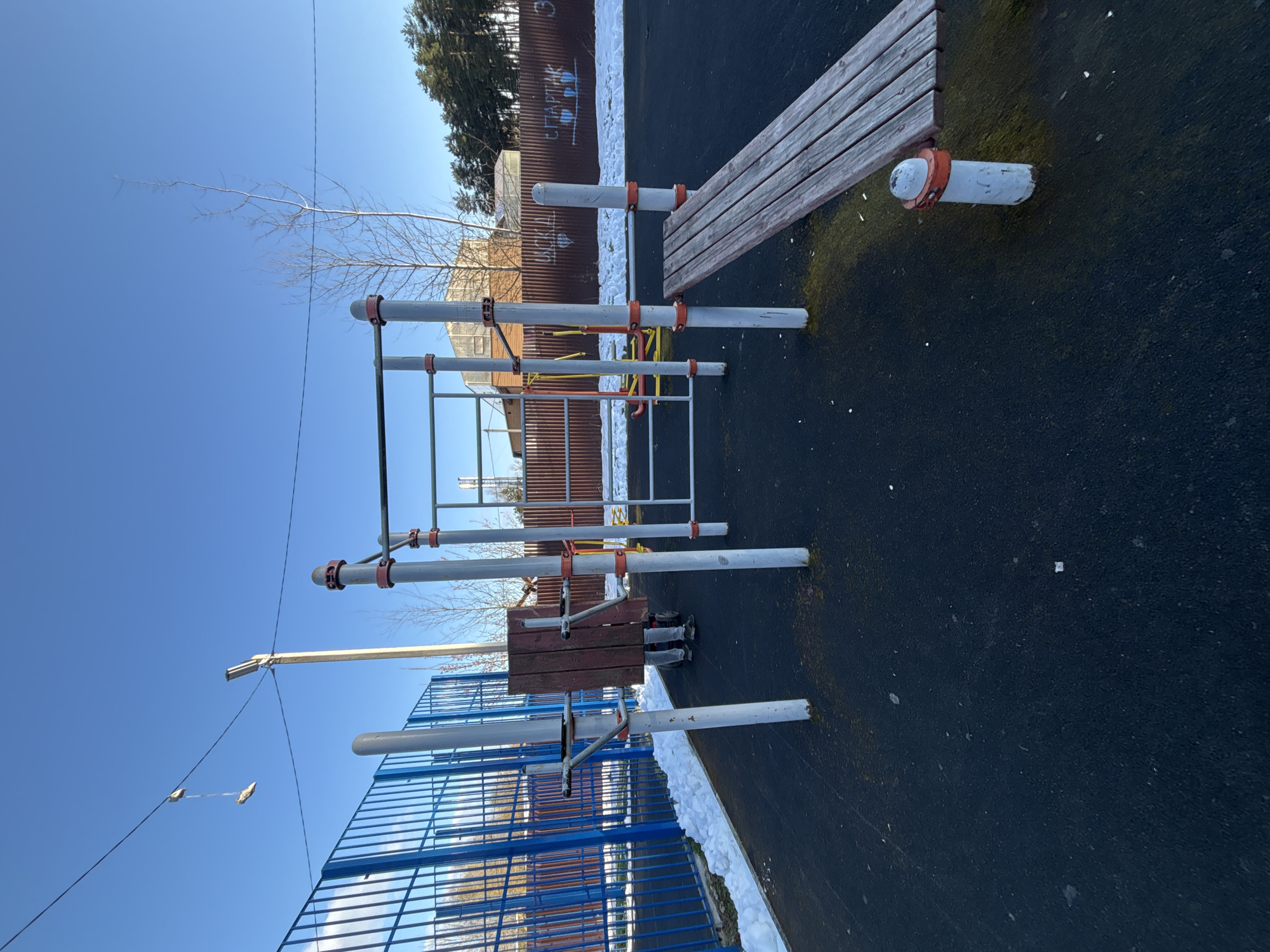 The image shows an outdoor fitness area in Razdolye, Russia. There is a blue metal fence, a climbing frame with gray metal poles and red connectors, and a wooden bench. The ground is black asphalt. Patches of snow are visible along the edges of the frame and fence. The background includes a metal fence with some graffiti on it and some trees. The sky is blue.