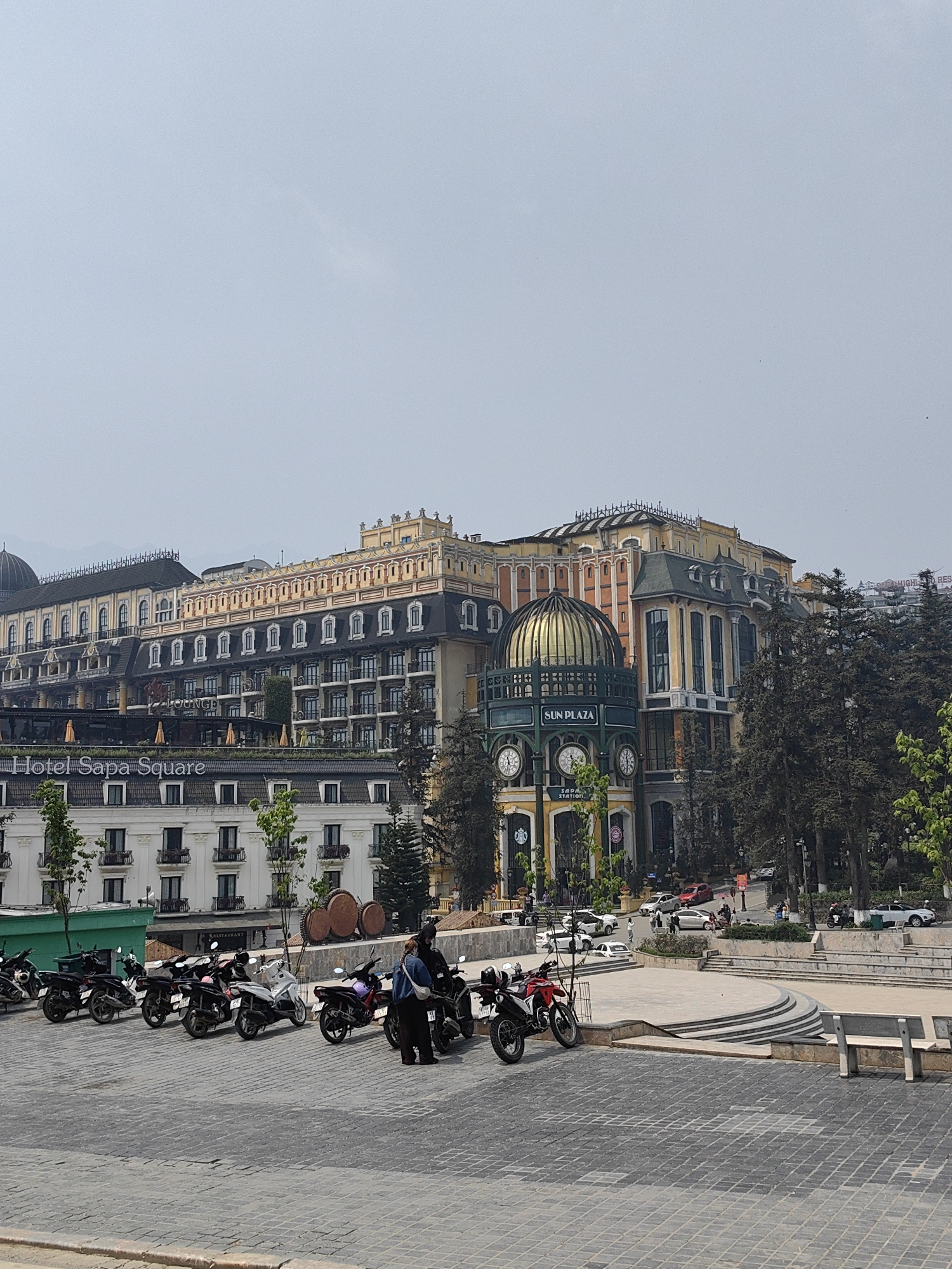 The image depicts a daytime street scene in Sa Pa, Vietnam, specifically within the town center near Sa Pa Square. The sky is uniformly light grey, indicating overcast conditions.
In the immediate foreground, a broad area of grey interlocking pavers constitutes the ground. To the left, a line of approximately eight parked motorcycles, mostly scooters of various colors (dark, white, red), is visible next to a low green structure. Slightly to the right of center in the mid-ground, two individuals are standing between two parked motorcycles; one person, wearing a light blue top and dark pants, faces away from the camera, interacting with a dark motorcycle, while the other is partially obscured.
The mid-ground features a multi-tiered plaza area constructed from light-colored stone, including curved steps and benches. Several tall, slender evergreen trees are dispersed throughout this area and behind the plaza.
The background is dominated by large, architecturally distinct buildings. On the left, the "Hotel Sapa Square" building rises multiple stories, characterized by its dark, sloping roofs, numerous white-framed dormer windows, balconies, and light-colored facades. To its right, the "Sun Plaza" building (identifiable by a "SUN PLAZA" sign and "SAPA STATION" below it) features a prominent golden dome with an intricate dark frame, multiple large clocks, and ornate, multi-colored facades (yellow, orange, green, grey). A Starbucks logo is visible on its ground level. Behind these main structures, additional buildings and distant mountains, partially obscured by haze, complete the scene. Several cars are visible on roads in the background, along with a few other pedestrians.
