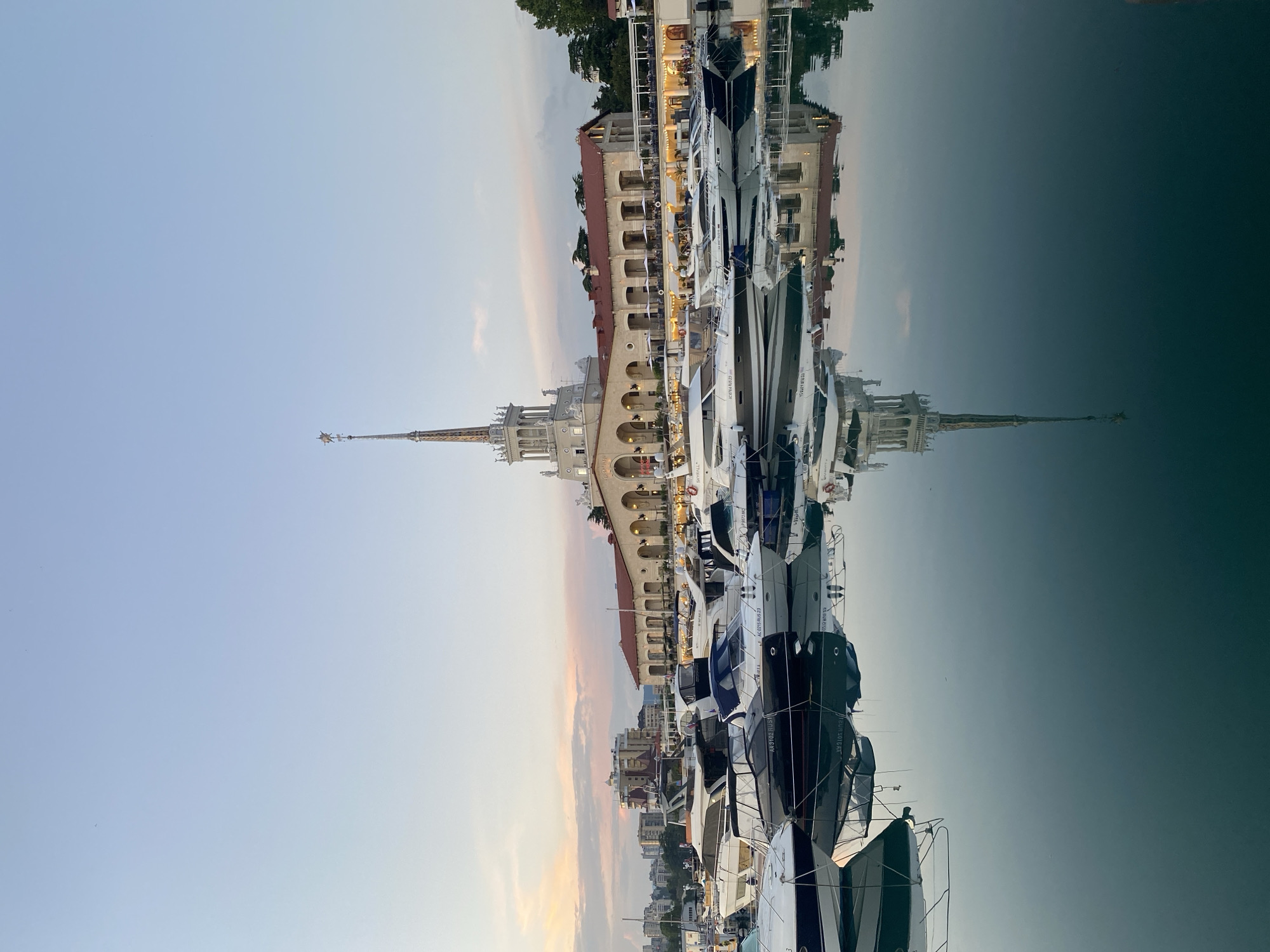 The image depicts the Sochi Sea Port in Sochi, Russia, at sunset. A large, ornate building with a tall spire dominates the background. Several yachts are docked in the foreground, their reflections visible on the calm water. The sky is a gradient of pale blue and pink, indicating the setting sun. The architecture of the building features arches, columns, and a prominent clock tower. The overall scene is one of tranquility and scenic beauty.