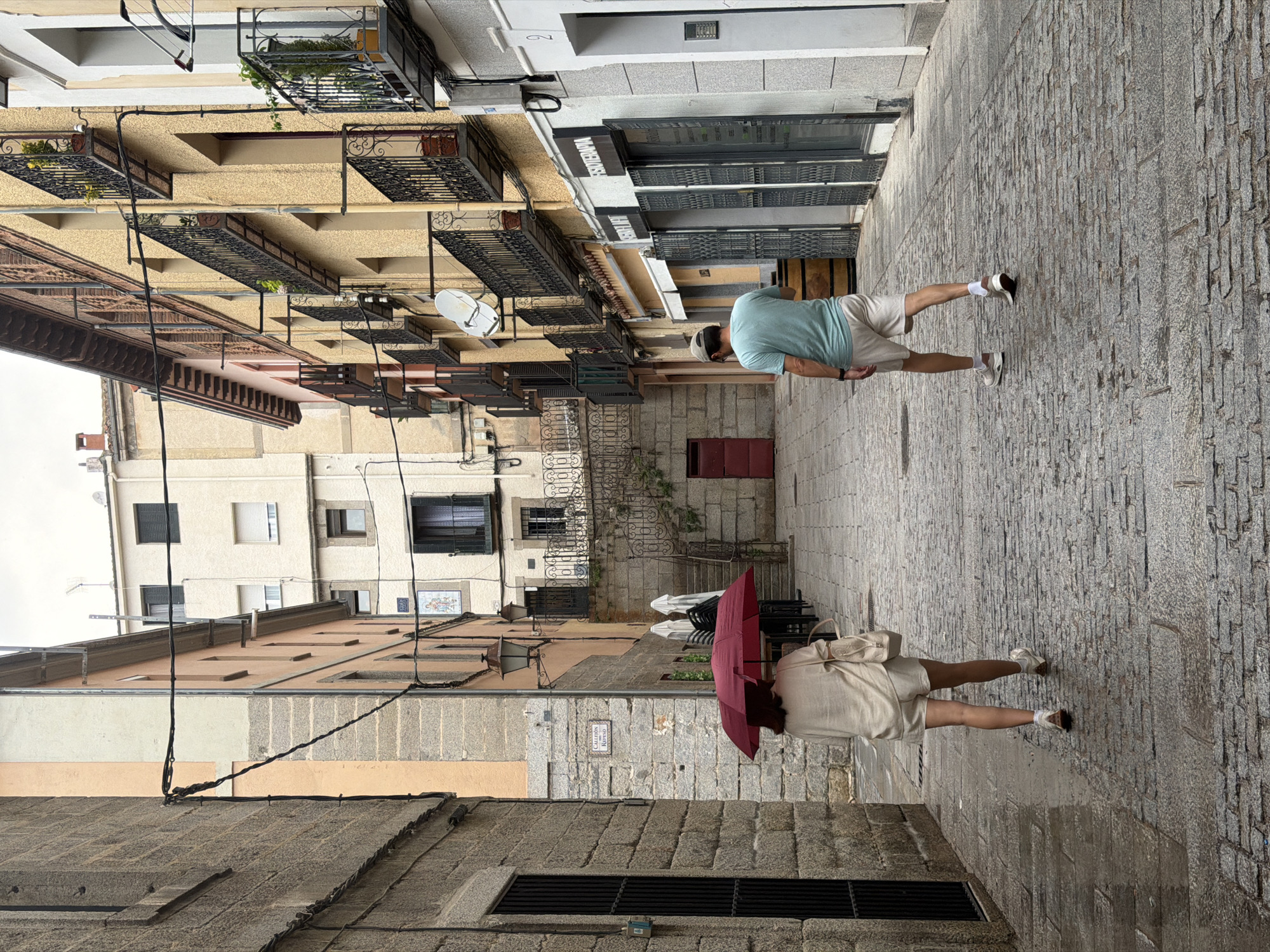 A man and a woman walk away from the camera down a narrow, cobblestone street in San Lorenzo de El Escorial, Spain. The street is lined with stone and stucco buildings. The woman, on the left, holds a red umbrella open above her. She wears a light-colored, loose-fitting top and matching shorts. The man, on the right, wears a light blue t-shirt and beige shorts. The sky is overcast, and the cobblestones appear wet, suggesting recent or ongoing rain. The buildings on the right have multiple balconies with black wrought-iron railings. Shops with closed metal shutters are visible on the ground floor of the building on the right. A red door is visible on the left side of the street further down.