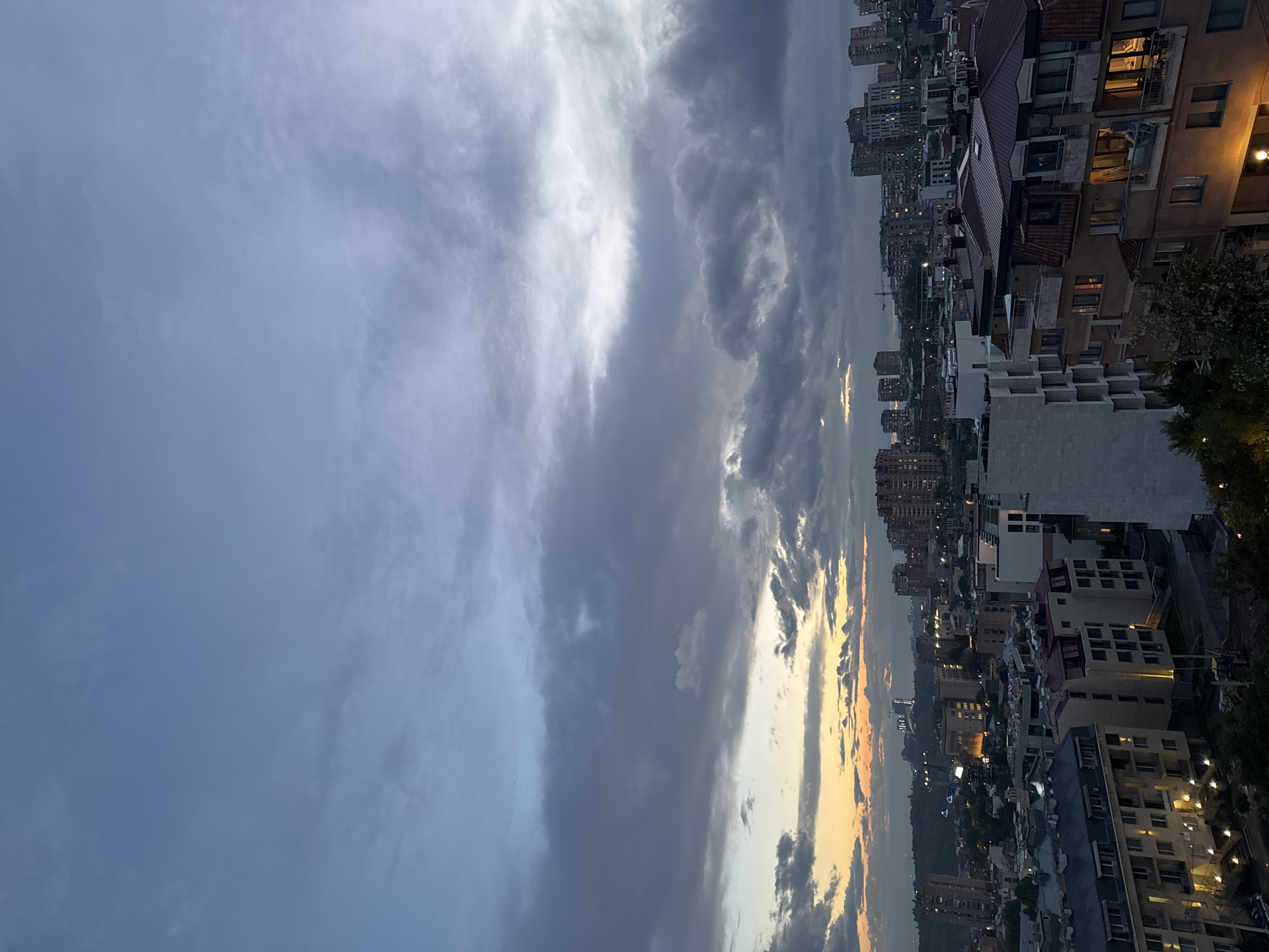 The image shows a view of Yerevan, Armenia, at dusk. A large portion of the frame is occupied by a cloudy sky with varying shades of gray and hints of orange near the horizon. Below the sky, a cityscape is visible with numerous buildings of varying heights and architectural styles. Some buildings have lights on in their windows, indicating it is evening. A construction crane is visible among the buildings in the distance. The scene appears to be photographed from an elevated perspective.