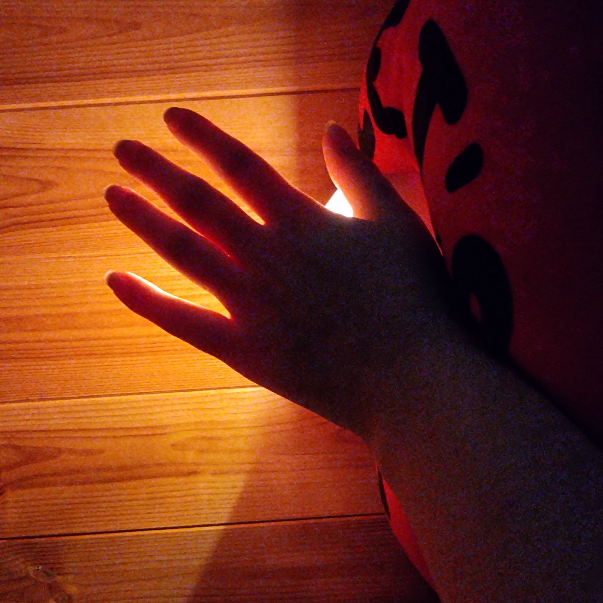 A person's hand is illuminated by a warm, orange light against a wooden plank background. The hand is positioned with fingers spread, casting a shadow. Below the hand, a portion of a red object with black patterns is visible. The lighting suggests an indoor setting, possibly a room with wooden walls or paneling. No specific location in Dunnae-myeon, South Korea can be determined from the image.