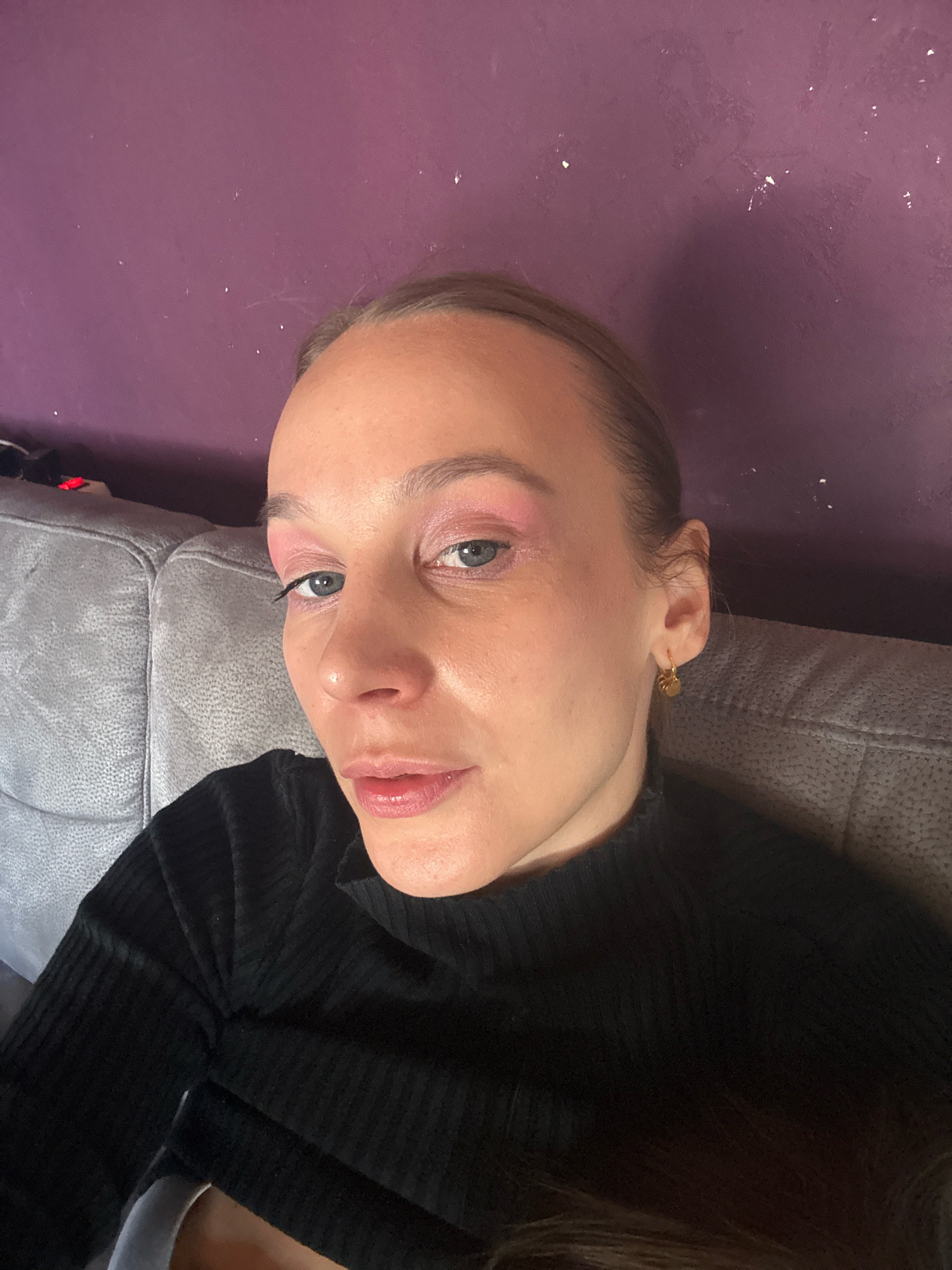 The image shows a close-up, selfie-style portrait of a woman with light skin and light brown hair pulled back. She is wearing a black, high-necked, ribbed sweater and a small gold earring with a circular charm on her right ear. Her eyes are open and looking towards the camera, and she is wearing pink eyeshadow and mascara. Her lips are slightly parted and appear to have a reddish-pink lipstick on. She is positioned on a grey sofa, which is slightly out of focus in the background. Behind the sofa is a textured purple wall. The overall lighting is dim, with some light sources creating reflections on her face and the wall. The scene is indoors, likely in a residential setting. There are no overt signs indicating a specific location within Überherrn, Germany.