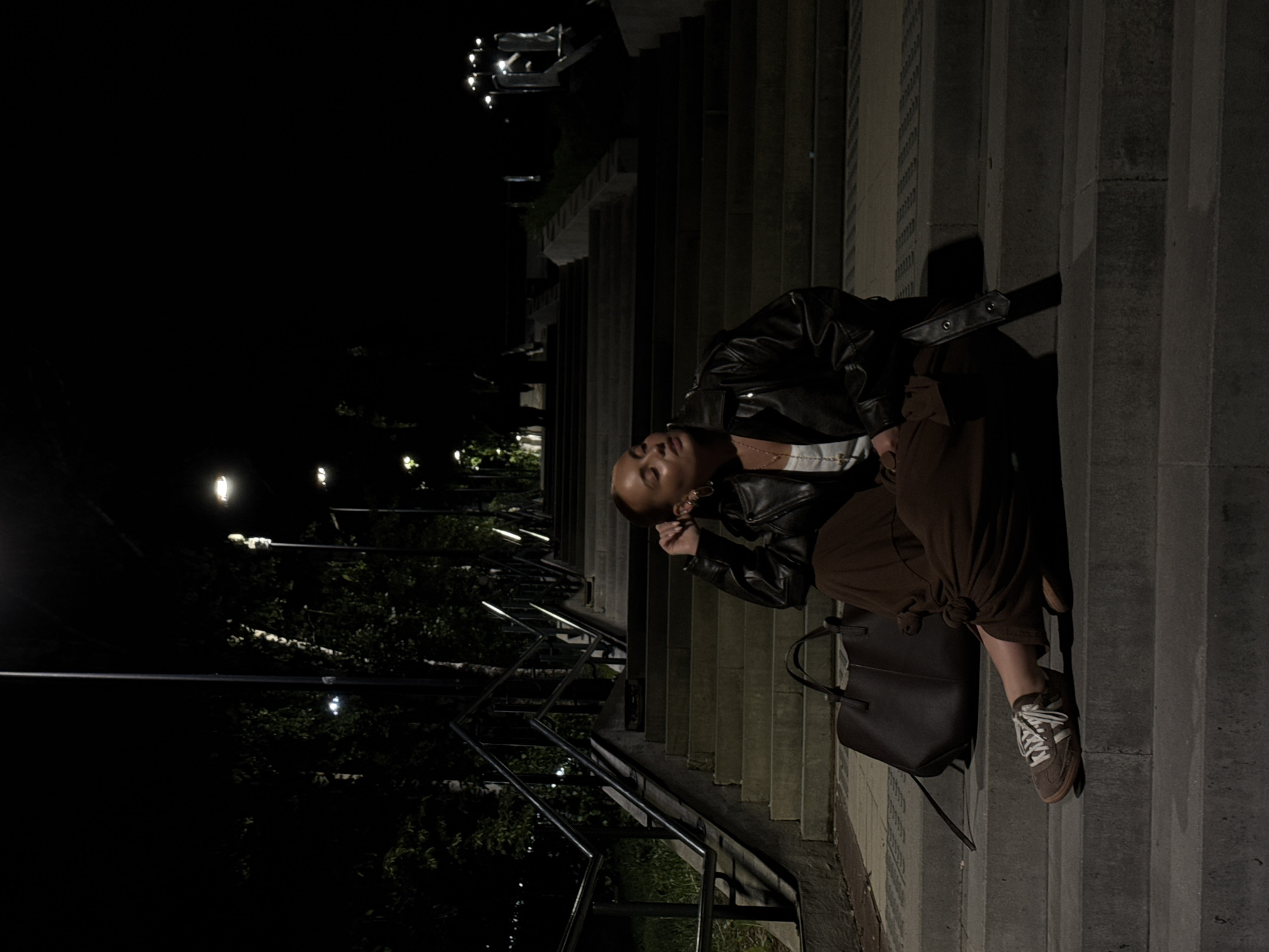 A woman sits on the steps of an outdoor staircase at night in Izhevsk, Russia. She wears a black leather jacket, brown pants, and brown and white sneakers. A dark brown tote bag sits on the steps next to her. The staircase is lined with metal railings, and the area is lit by streetlights. Beyond the staircase, there is foliage and more streetlights. The image is dark and grainy.