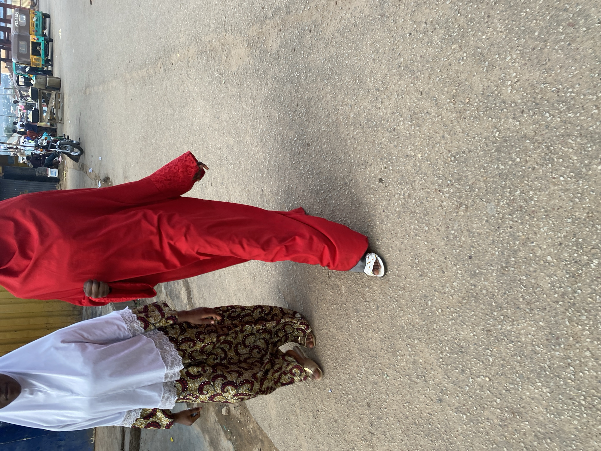Two individuals walk down a paved road in Jos, Nigeria. The person on the left wears a white hijab and a patterned skirt, while the person on the right is dressed in a flowing red garment. In the background, a motorcycle is parked, and several small vehicles, including a green and brown auto rickshaw, are visible on the road. Structures and market stalls line the left side of the street, suggesting a busy commercial area. The ground is asphalt with visible aggregate.