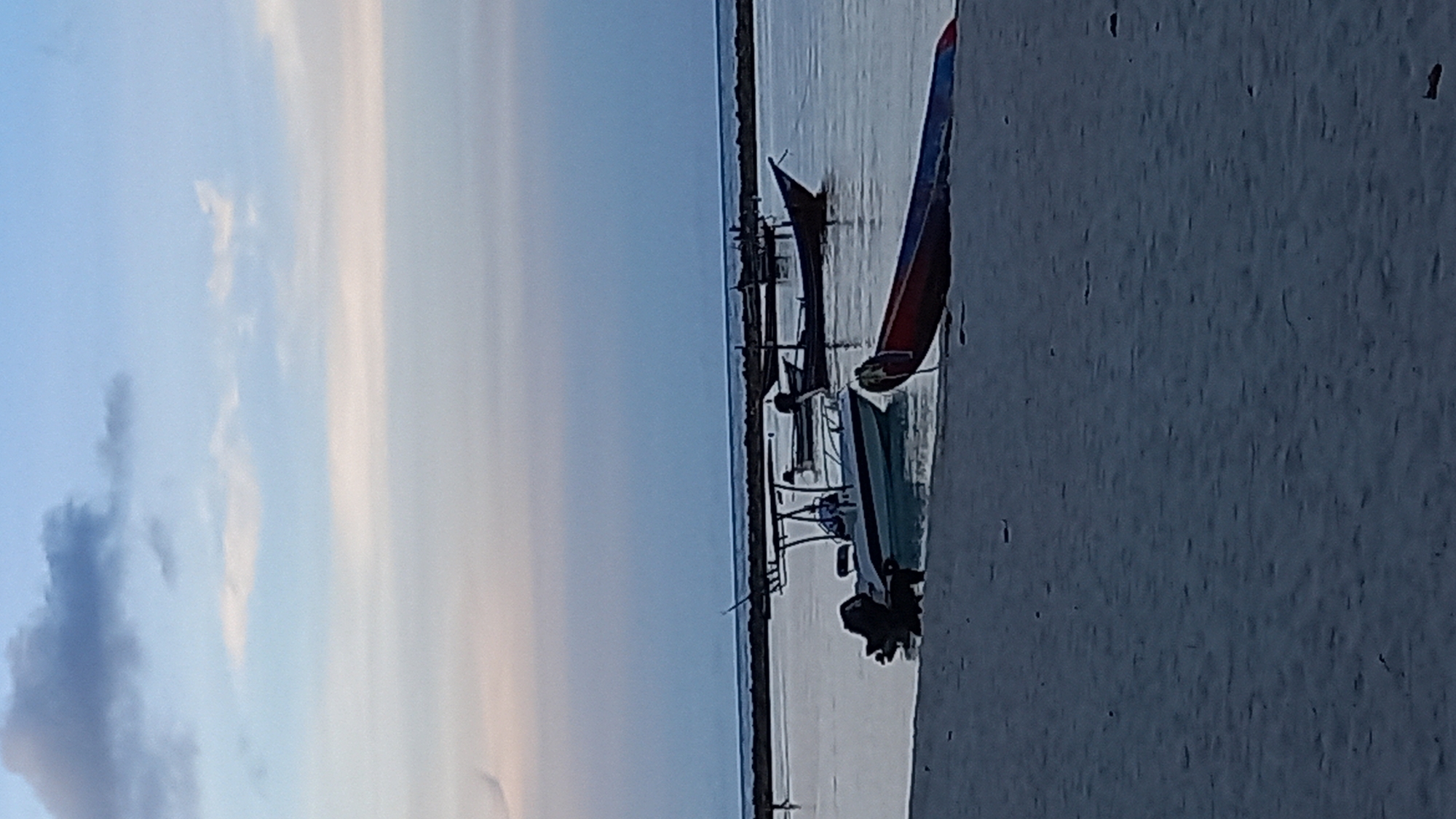 A serene beach scene at twilight in Koh Phangan, Thailand, features a calm body of water reflecting the soft hues of the sky, transitioning from pale blue to a delicate pinkish-grey. Several boats are visible, some gently bobbing on the water and others resting on the sandy shore. In the foreground, a long, narrow red and blue boat lies partially on the beach, its hull angled towards the water. Slightly further out, a white motorboat with a canopy and an outboard motor is moored. Behind it, several traditional wooden fishing boats are also anchored, their dark silhouettes contrasting with the lighter water. A low, dark horizon line, possibly a breakwater or a distant landmass, separates the water from the sky. The beach itself is composed of fine, grey sand with small debris scattered across its surface. The sky is dotted with scattered clouds, one dark grey cloud formation is visible towards the top left of the frame. The overall impression is one of peaceful tranquility.