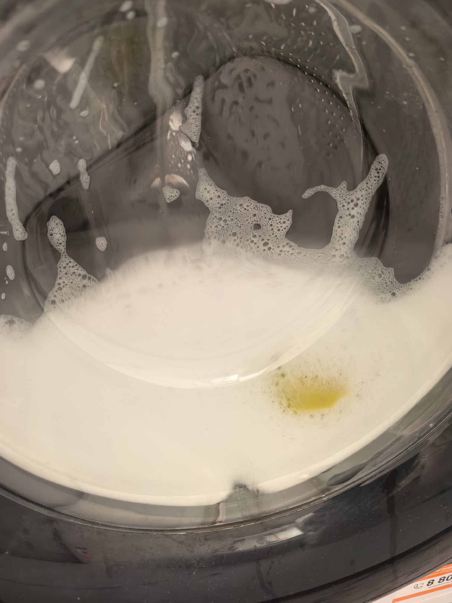 The image displays the interior of a front-loading washing machine, viewed through its transparent circular door. The machine is actively engaged in a wash cycle, indicated by the considerable amount of white, frothy foam and soapy water filling the bottom half of the drum. Foam also adheres to the inner surface of the glass door.
A distinct, bright yellow liquid or substance is visible within the white foam, concentrated towards the bottom right area of the visible drum interior. This yellow substance contrasts sharply with the surrounding white suds. The interior of the washing machine drum, visible through the water and foam, appears to be dark grey or black.
In the bottom right corner of the original image, a sliver of an orange label or tag is partially visible. This label features Cyrillic text. Recognizable fragments include "ТРИН", "СЕР", and a prominent "8 80" followed by further Cyrillic characters, which suggest a Russian toll-free phone number (e.g., "8 800"). Below this, more text reads "позвоните по" (call by), followed by additional numbers. These textual cues are consistent with the provided location context of Sterlitamak, Russia. The image captures the washing machine in what appears to be an indoor setting, likely a laundry area. There are no people visible, and no specific time of day or emotional context can be discerned.