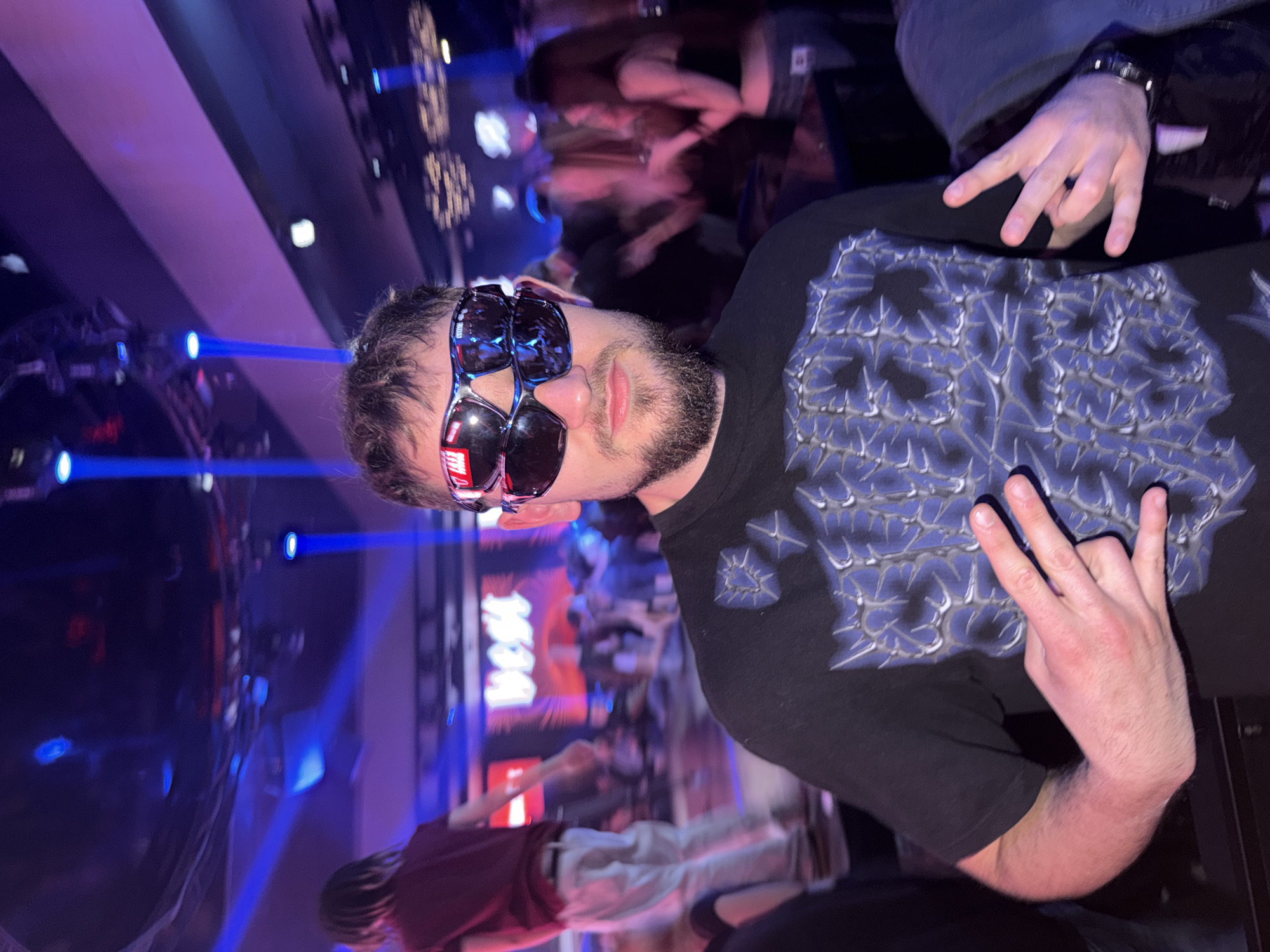 This image, rotated 90 degrees counter-clockwise, depicts a lively indoor event, likely a concert or club, set in Plovdiv, Bulgaria. The foreground is dominated by a young man with a beard, wearing a dark t-shirt with a prominent blue-grey spiky or thorny graphic design. He is wearing two pairs of reflective sunglasses stacked on his face; text like "SELLAS" is visible on one lens, while "HELLAS" or similar text might be on the other. He wears a dark watch on his left wrist.
The man poses confidently for the camera, making a "rock and roll" or "devil horns" hand gesture with his right hand, and a similar gesture with his partially visible left hand. The background is a blur of motion, indicating a crowd of people enjoying the event. The venue is illuminated by striking blue and purple stage lights, casting dynamic beams across the dark space and reflecting off surfaces. On a screen or sign in the mid-background, the word "BEAT" is clearly visible in white/red against a dark background, reinforcing the musical or entertainment context of the scene. The overall ambiance is energetic and characteristic of a late-night social gathering.