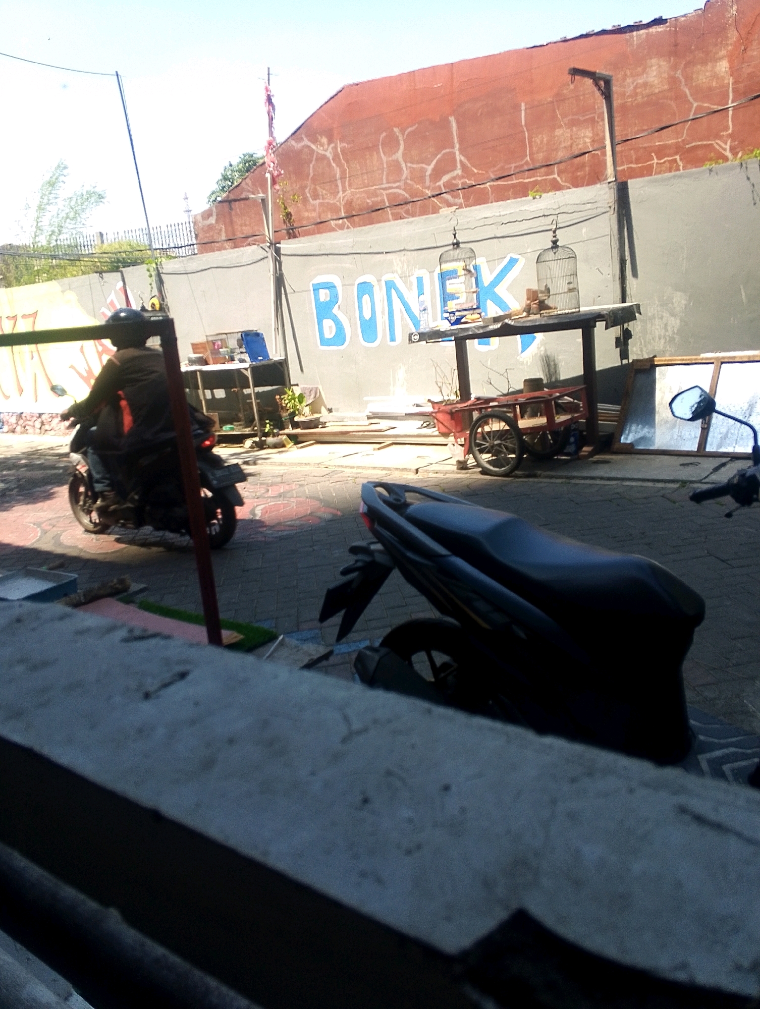 A medium shot captures a street scene under clear daylight in an urban environment, possibly a market or roadside area. The foreground is dominated by the edge of a textured concrete structure, casting a shadow and creating a barrier. To the left, a portion of a patterned, possibly tiled, ground is visible.
In the mid-ground, a dark-colored motorcycle is parked and partially obscured by the foreground element. Further back, a person wearing a helmet and dark clothing rides another motorcycle, their body angled away from the camera, suggesting forward motion. This motorcycle is also dark-colored.
Behind the moving motorcycle, a low, gray wall runs horizontally across the scene, painted with large, blue, block letters that spell "BONEK". Above the wall, a reddish-brown, textured wall with white, crack-like markings creates the upper background. A simple, dark-colored table with a dark surface stands in front of the gray wall, supporting two birdcages, one with a bird inside. A small, red cart with two wheels and a wooden bed is situated to the right of the table. Several framed objects, possibly mirrors or windows, are leaning against the gray wall to the right of the table.
A thin, black cable stretches diagonally across the upper left portion of the image, anchored to a tall, thin pole with a red and white banner attached to its top. In the far background, beyond the red wall, hints of greenery and structures are visible, along with a clear, pale blue sky. The overall impression is one of a bustling, perhaps working-class, urban setting.