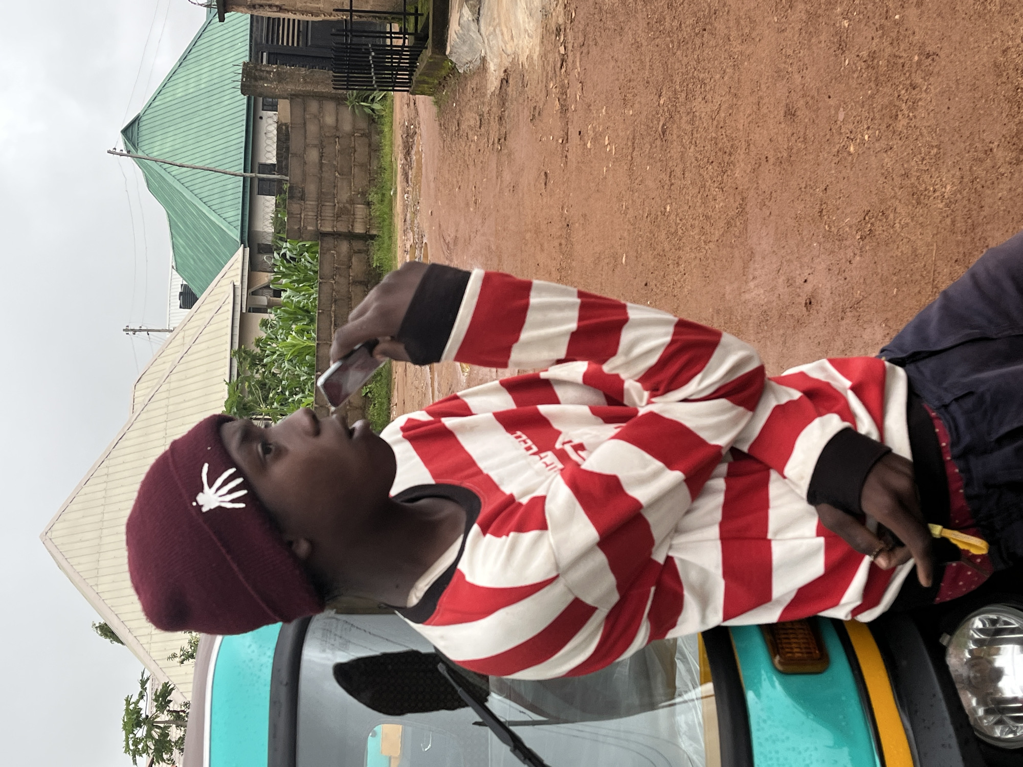 In Barakin Akawo, Nigeria, a young Black male wearing a maroon beanie with a white spider emblem and a red and white striped long-sleeved shirt stands next to a teal and yellow auto-rickshaw. He is holding a silver mobile phone to his mouth as if speaking or about to speak. His left hand is resting on the auto-rickshaw, and his right hand is near his mouth. The ground is a reddish-brown dirt road with scattered puddles, indicating recent rain. In the background, several buildings with distinct rooflines, including a prominent green gabled roof and a light-colored pyramidal roof, are visible. A brick wall and a black metal gate line the right side of the road. The sky is overcast.