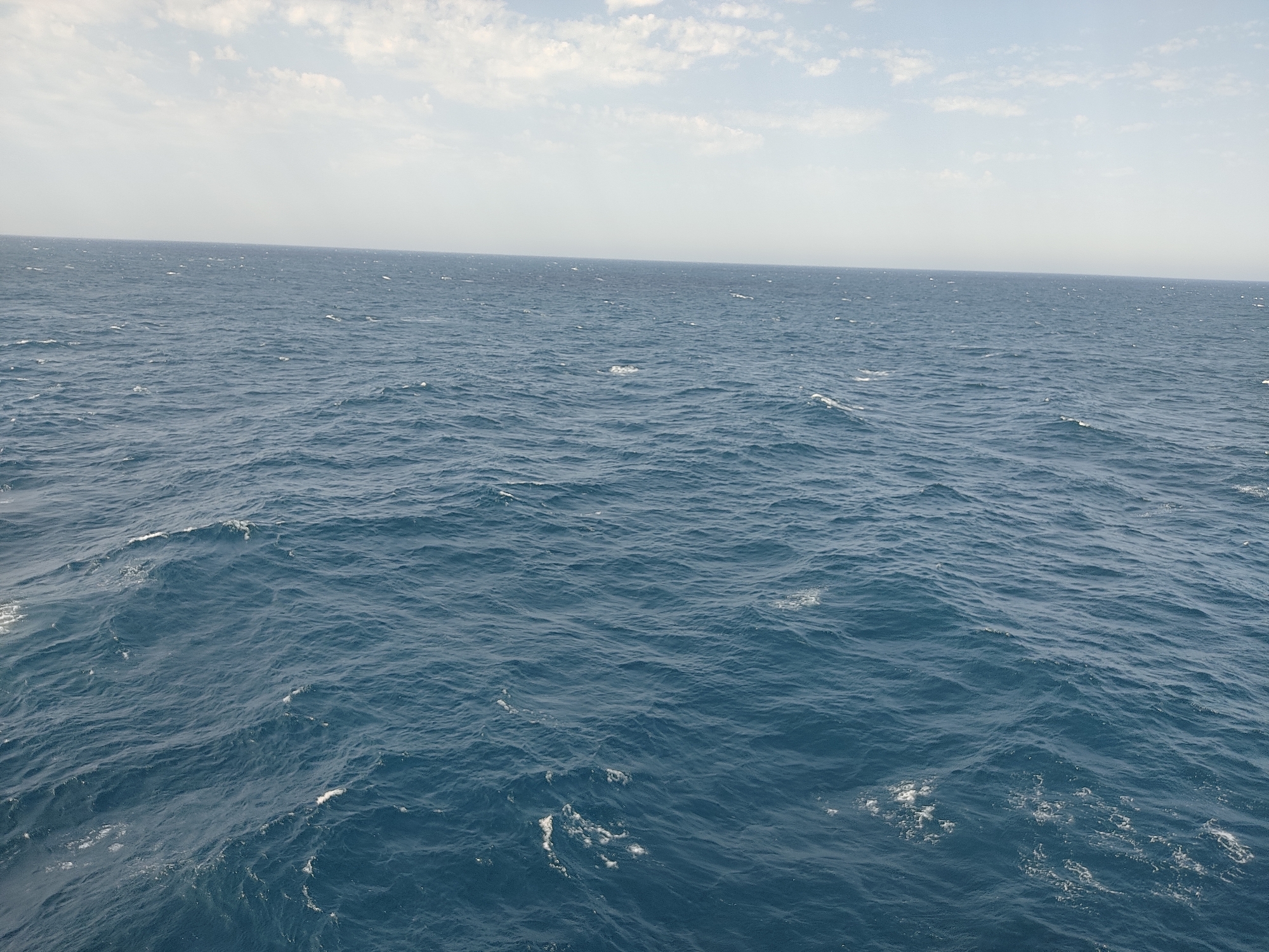 The image shows a wide expanse of the ocean under a hazy sky. The water is a deep blue color with small whitecaps and ripples on its surface, indicating some wave activity. The horizon is visible as a faint line where the sea meets the sky. The sky is a pale blue with scattered, wispy white clouds, particularly in the upper portion of the frame. There are no visible landmasses, boats, or any other objects in the image. The scene is an unobstructed view of the open sea.