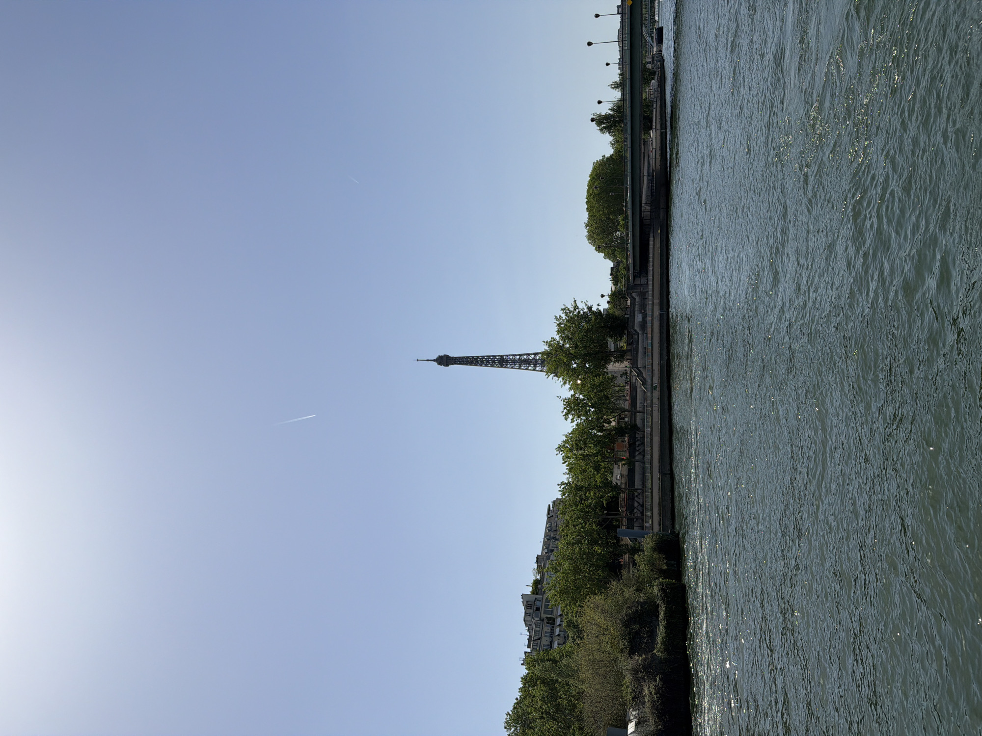 The image shows the Eiffel Tower in Paris, France, from a low vantage point, likely from the Seine River. The tower is visible in the center of the frame, piercing the clear blue sky. A jet stream is slightly visible to the left of the tower. The foreground shows the shoreline with buildings and trees. The Seine River is on the right. The image is rotated by 90 degrees clockwise.