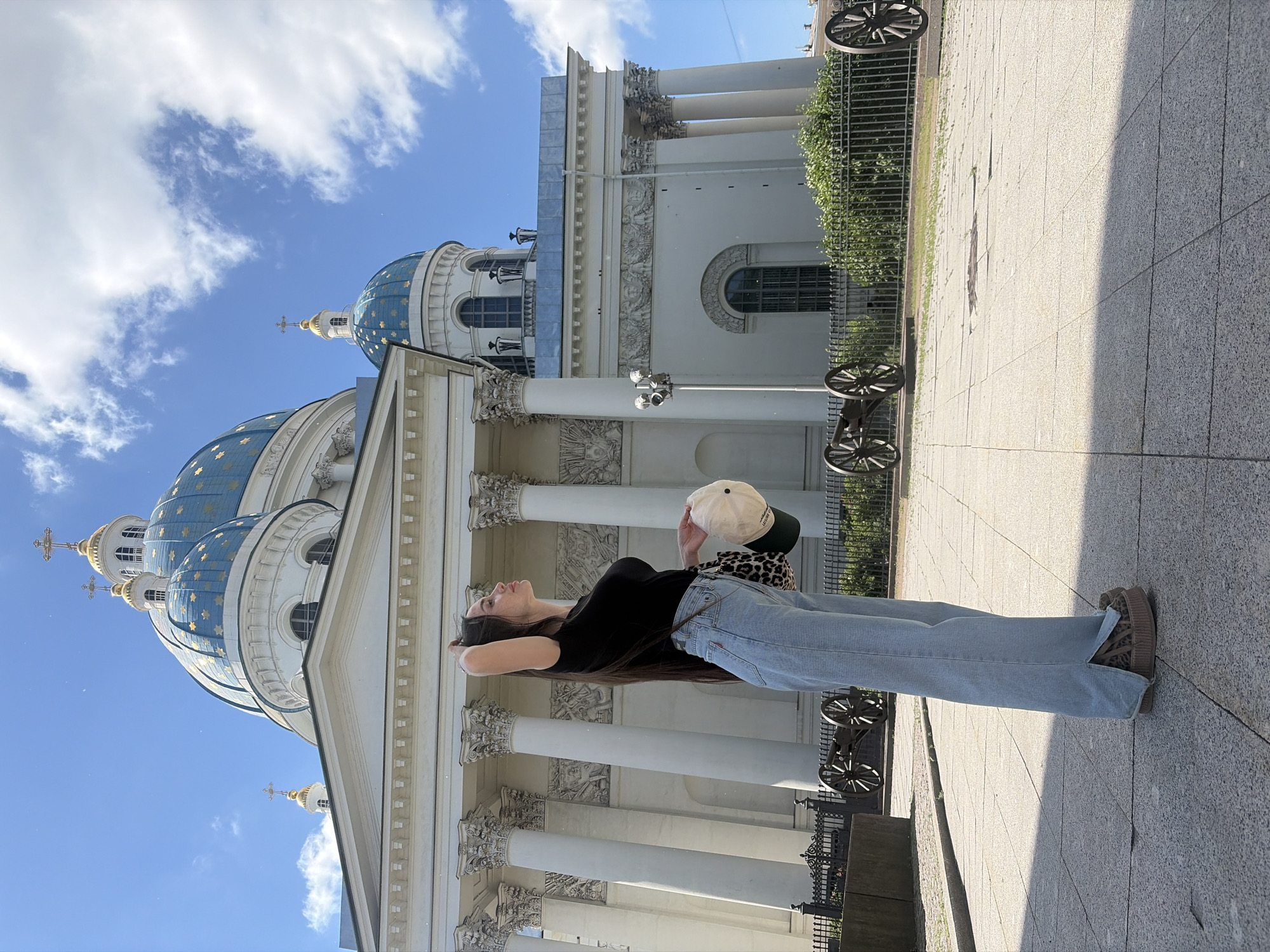 A young woman with long dark hair is standing in front of the Trinity Cathedral, a prominent landmark in Saint Petersburg, Russia. She is wearing a black t-shirt, light blue wide-leg jeans, and leopard-print platform shoes. She is holding a beige baseball cap and a leopard-print bag in her hands. The cathedral has a white facade with large columns and blue domes topped with golden crosses. In the foreground, there are several historical cannons on display, enclosed by a black metal fence. The sky is blue with scattered white clouds, and the ground is paved with gray tiles, casting shadows from the building and other elements. The overall scene depicts a sunny day in the city.