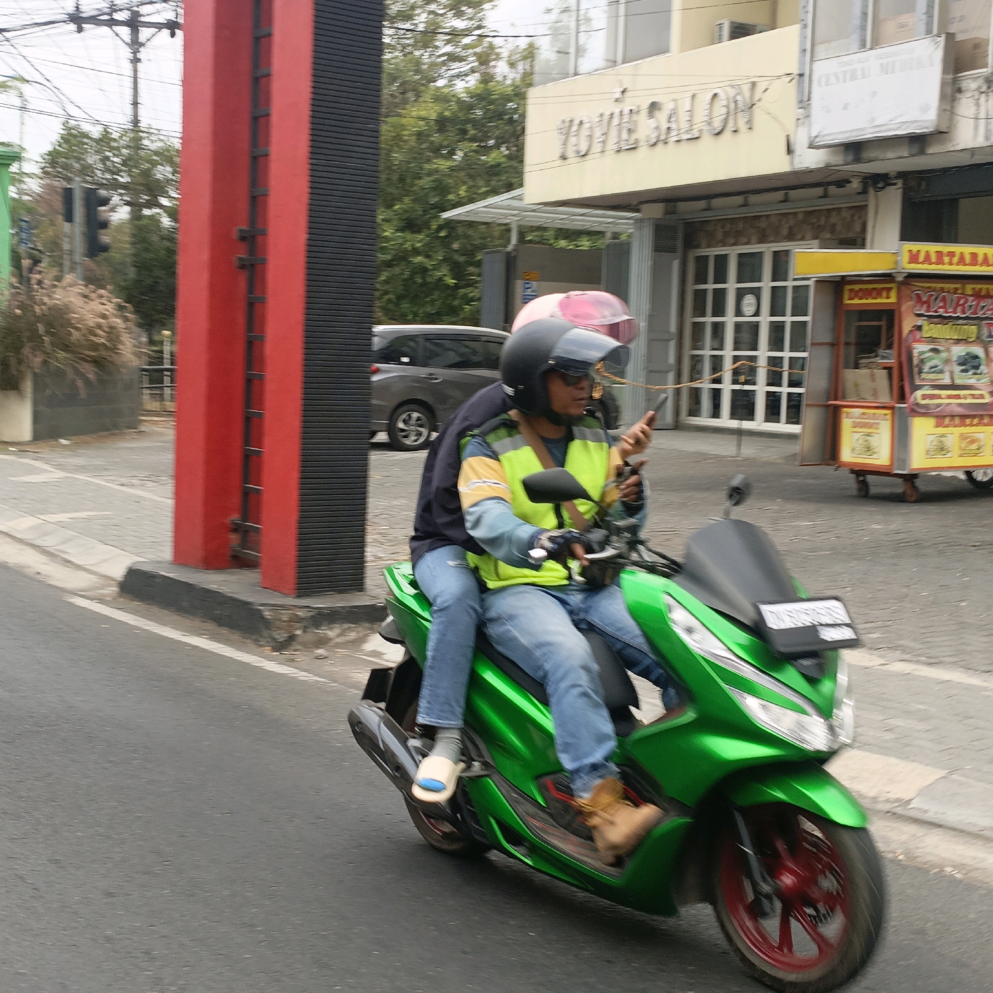 Two individuals are riding a green scooter on a street in Salatiga, Indonesia. The person in the front is wearing a black helmet, sunglasses, a yellow and green reflective vest over a blue long-sleeved shirt, and blue jeans. They are holding a smartphone in their right hand while operating the scooter. The passenger behind them is wearing a dark blue jacket over a lighter colored shirt and blue jeans. Their left arm is around the driver. They appear to be wearing slippers. The scooter has a visible license plate. In the background, a red and black pillar is present, along with trees and buildings. One building has signage that reads "YOYIE SALON". Another structure appears to be a food stall with colorful signage. A grey car is visible in the distance. The road surface is asphalt with a white painted line.