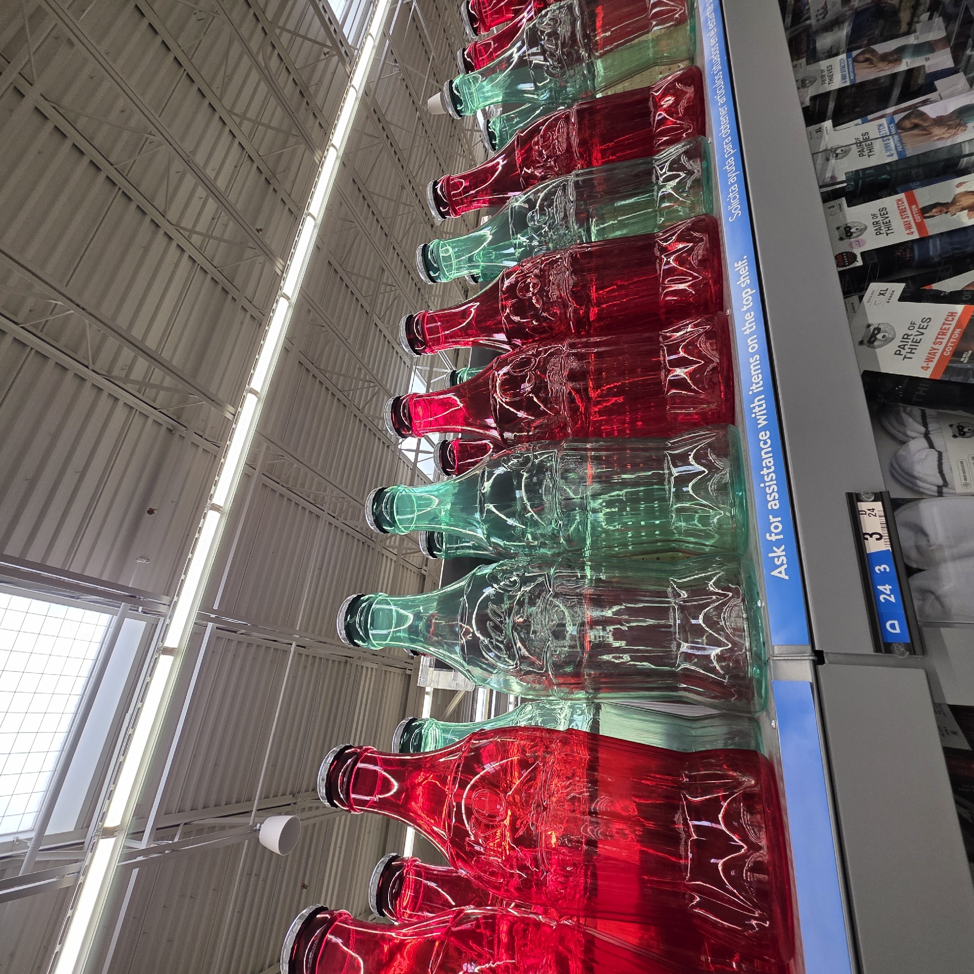 The image shows a shelf in a retail store with rows of large, decorative Coca-Cola bottles, alternating in red and green. The bottles are made of glass and have the iconic Coca-Cola logo embossed on them. The bottles are filled with liquid, the red ones with a red liquid and the green ones with a green liquid. Some of the bottles have silver caps. Above the bottles, a white sign with blue text reads "Ask for assistance with items on the top shelf." Below the bottles, another shelf displays packages of socks and underwear. The ceiling of the store is visible in the background, with fluorescent lighting and exposed metal beams. The image is taken from a low angle, looking up at the bottles. The location is a retail store in Tigard, Oregon, United States of America.