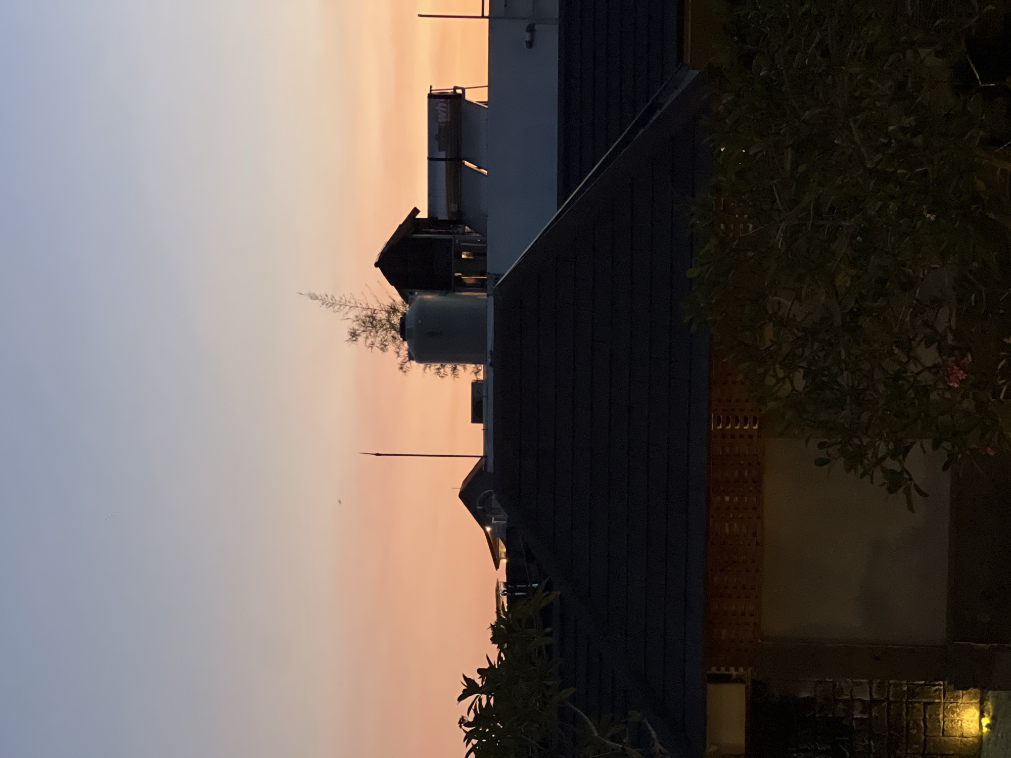 The image captures a twilight cityscape in North Kuta, Indonesia. The sky is a gradient of pale blue to orange, indicative of sunset or sunrise. The foreground is dominated by rooftops with dark tiles. A water tank and some type of utility structure are visible atop one of the buildings. Vegetation, possibly trees or bushes, frames the lower and right edges of the image. The overall lighting suggests it is either dusk or dawn. There is no evidence of human presence or activity in the frame.