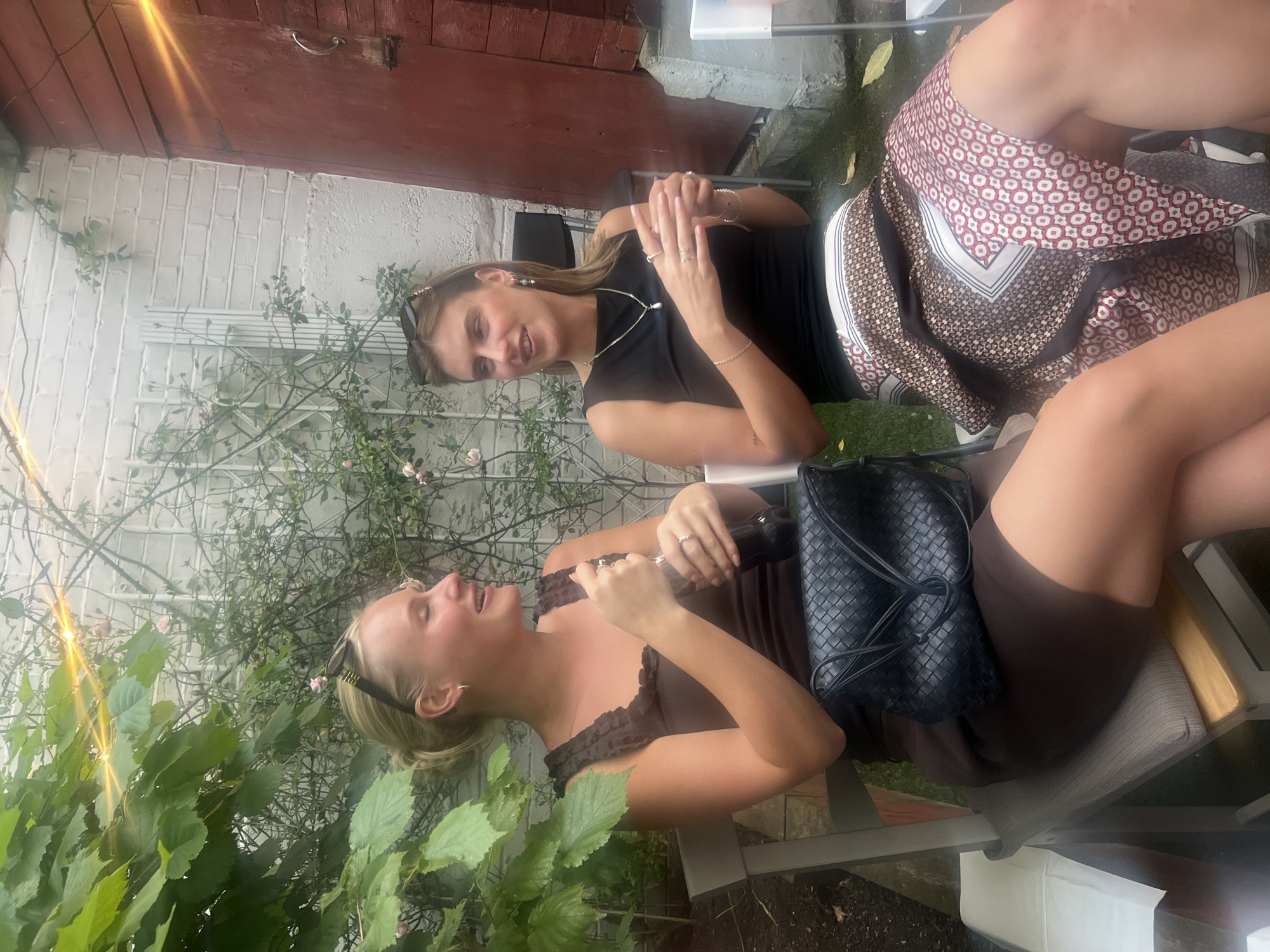Two women are seated outdoors at a table. One woman is wearing a black tank top, a silver necklace, a brown and patterned scarf, and sunglasses are perched on her head. The other woman is wearing a brown ruffled dress. A black purse sits on the table near her. There is a brick wall and white wall nearby. The women are in an outdoor area surrounded by foliage. The location appears to be a garden or patio area, likely in London based on the user instructions. The women appear to be having a conversation.