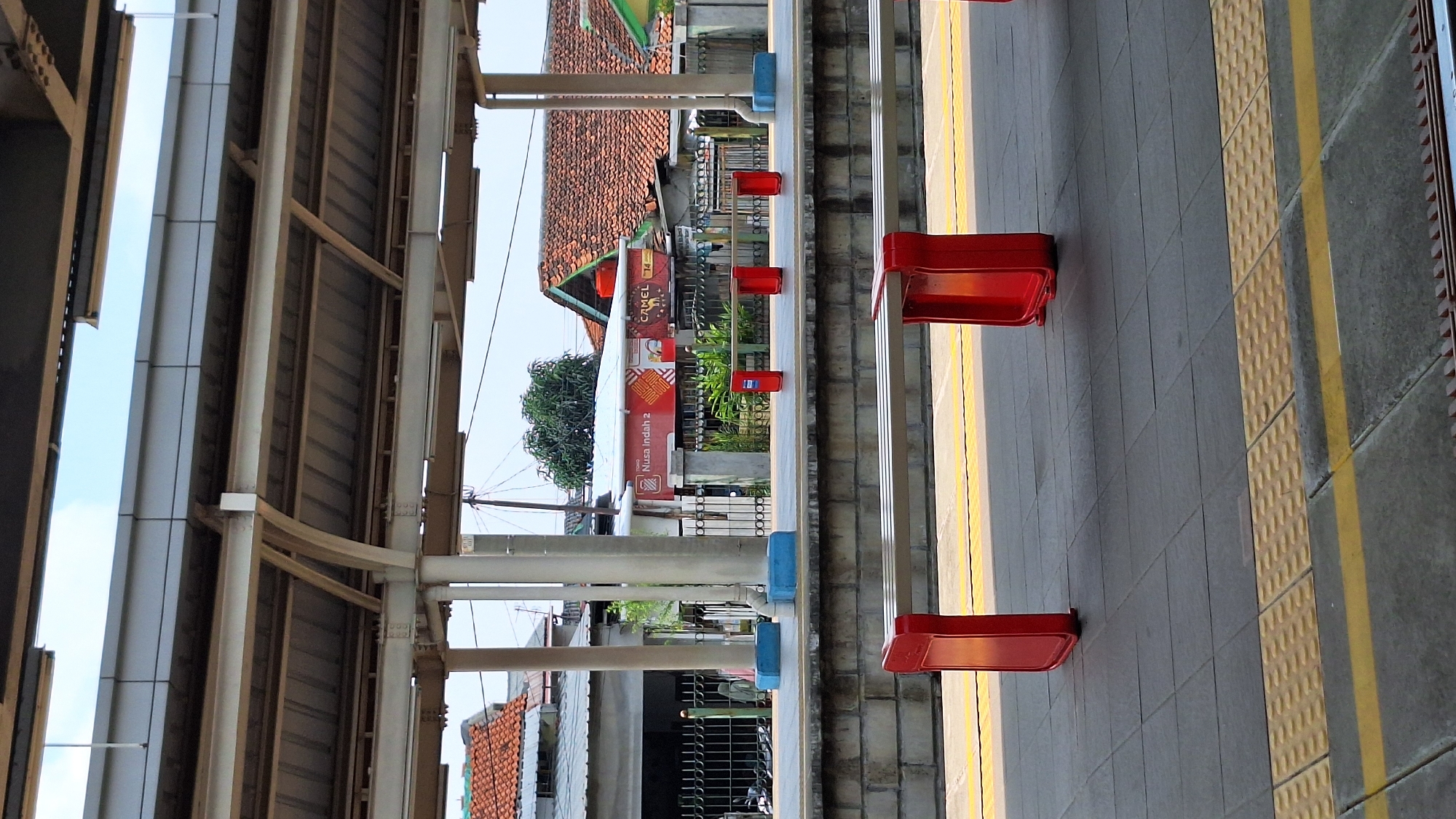 The image depicts a train platform in Tambun, Indonesia. The platform is constructed with gray tiles and features yellow tactile paving along the edge, indicating a safety zone. Red, cantilevered benches are positioned along the platform. Overhead, a metal structure supports the platform's roof, with corrugated metal panels visible. Across the tracks, a view of buildings with red tile roofs and various advertisements are visible. A sign reading "Nusa Indah 2" can be discerned. The sky is partially visible between the buildings and the roof structure.
