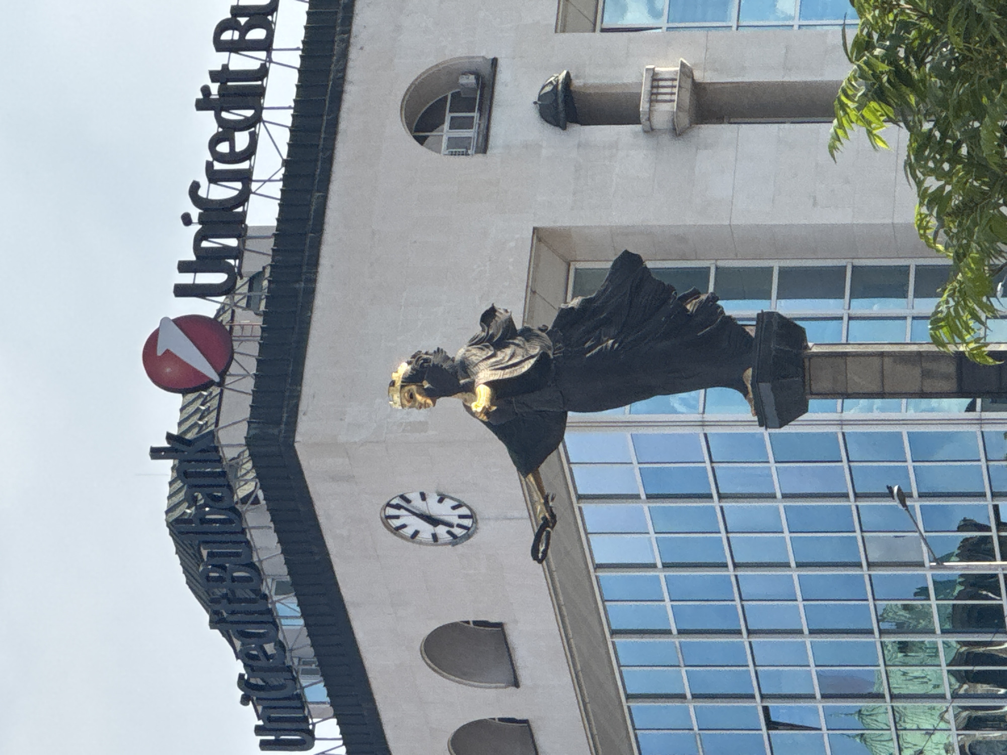 The image, rotated 90 degrees clockwise for proper viewing, depicts a modern building in Sofia, Bulgaria, featuring a large statue and the prominent "UniCredit Bulbank" sign.
The central subject is a dark-colored, majestic statue of what appears to be an allegorical or mythical figure, possibly female, with striking golden details on its face and head. The figure is shown in motion, with drapery flowing around it, and holds a dark ring or wreath in one hand, extending outwards from the building's facade.
Below the statue, on the building's light-colored facade, there is a circular analog clock with a white face and black hands and markings. The building itself is multi-story, characterized by large, rectangular, blue-tinted windows that reflect the bright sky and surrounding environment. Some smaller, rounded window openings are also visible on the upper levels.
Dominating the upper left portion of the building is a large, dark-lettered sign spelling "UniCredit Bulbank," with the bank's distinctive red circular logo featuring a white arrow positioned beneath the text.
The scene is set during the daytime, indicated by the bright, clear lighting and the reflections in the windows. A vibrant green tree is partially visible on the right side of the frame, adding a touch of nature to the urban setting. The overall impression is one of a prominent financial institution adorned with artistic elements, standing tall under a clear sky.