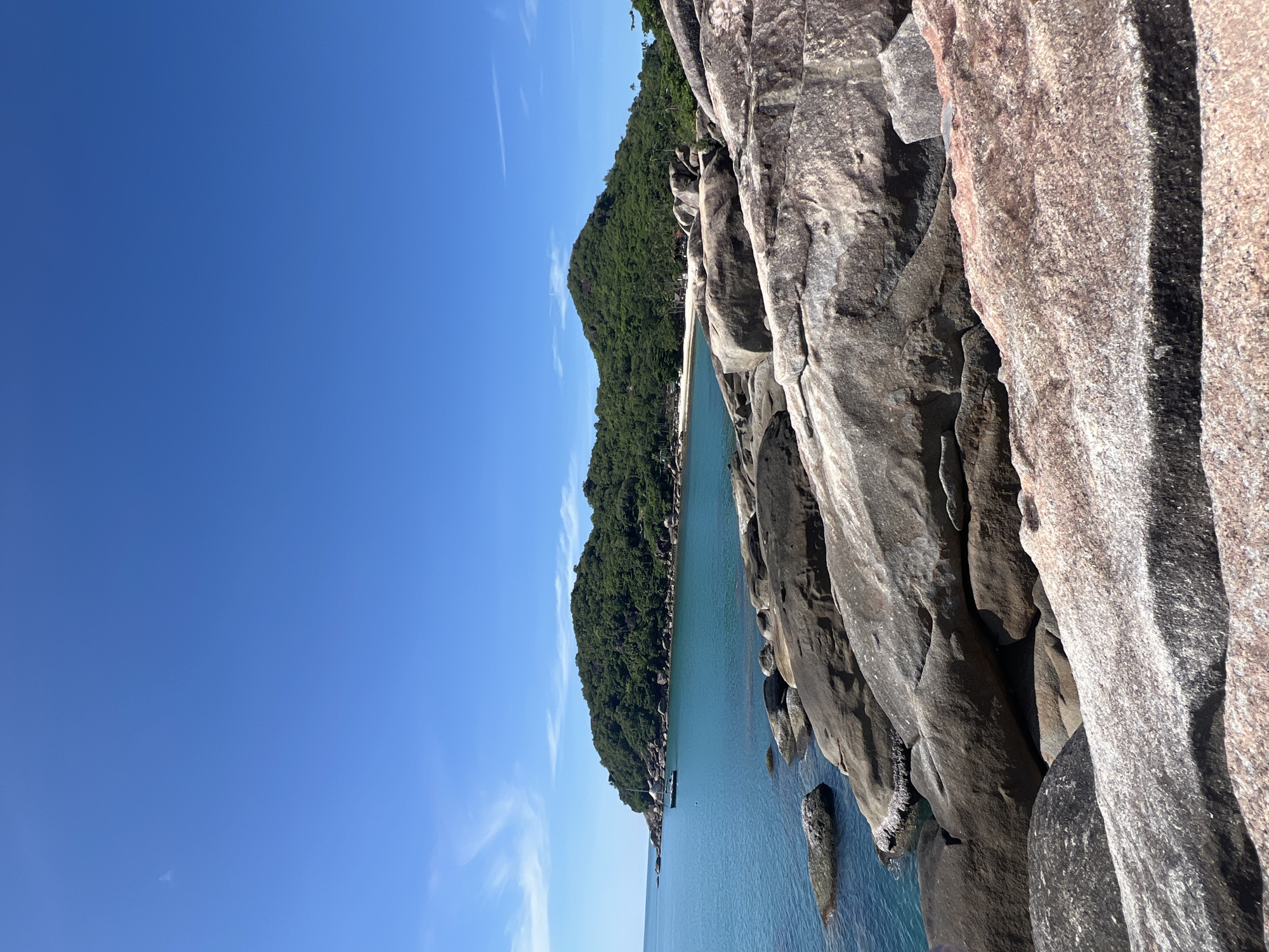 A wide shot captures a tropical coastal scene with a clear blue sky and lush green hills. In the foreground, large, weathered granite boulders dominate the landscape, their surfaces textured and varied in color from gray to reddish-brown. Some of these rocks are partially submerged in the turquoise water. The ocean stretches out to the left, calm and reflective, with a few smaller rocks visible near the shore. In the mid-ground, a sailboat with a white sail is anchored in the bay. The background features a densely vegetated hillside that slopes down to a sandy beach, suggesting a remote and serene location. The overall impression is one of natural beauty and tranquility.