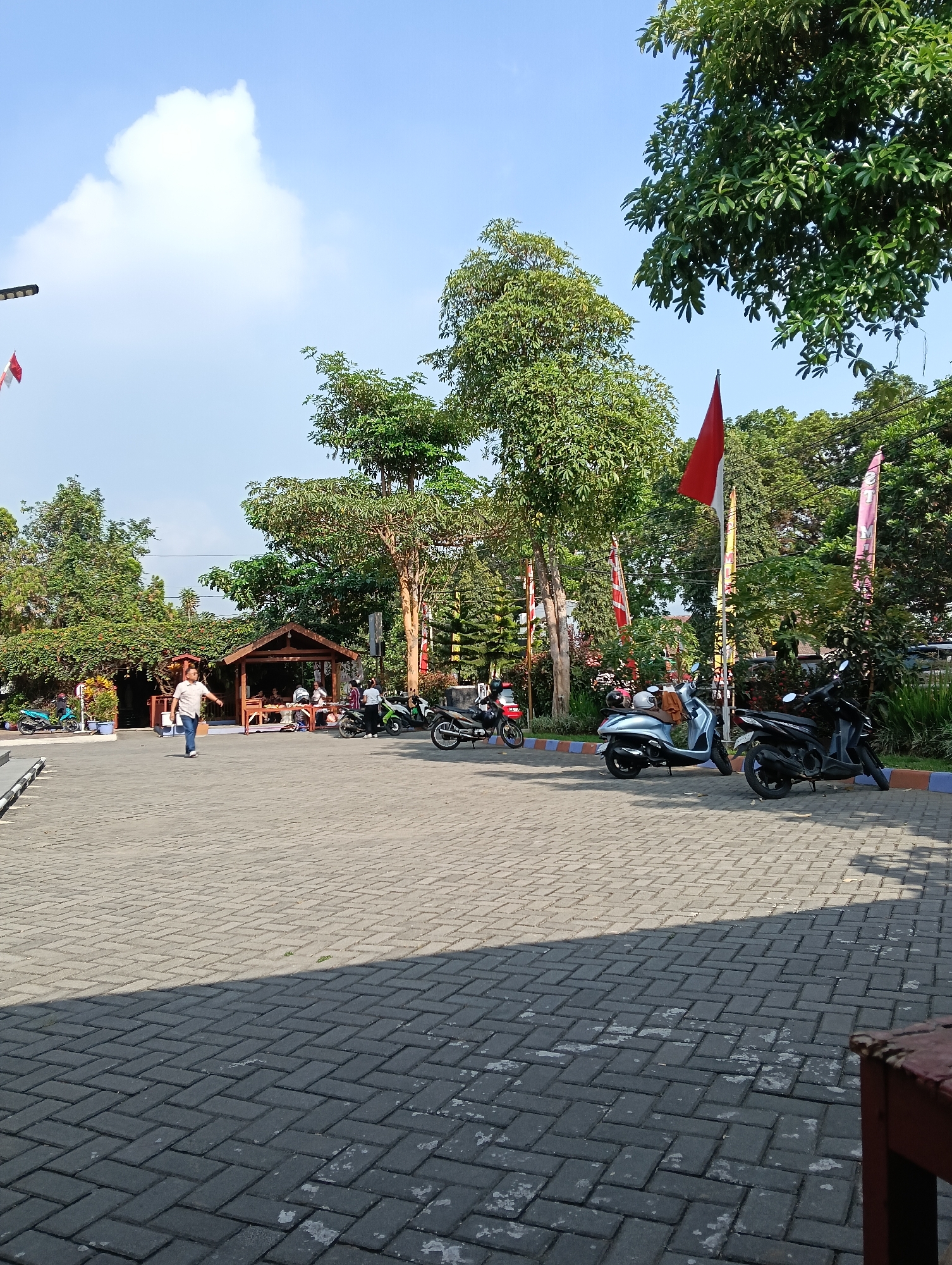 The image shows an outdoor scene in Banyurojo, Indonesia, on a sunny day. A paved area, composed of gray interlocking bricks, dominates the foreground, casting shadows. In the middle ground, a group of people are gathered near a wooden gazebo with a thatched roof. Several motorcycles are parked in front of the gazebo and to the right. Behind the gazebo, lush green trees and foliage create a backdrop, with some red and white flags displayed on poles. The sky is clear blue with a large white cloud visible on the left side. A street lamp with a light fixture is positioned on the far left.