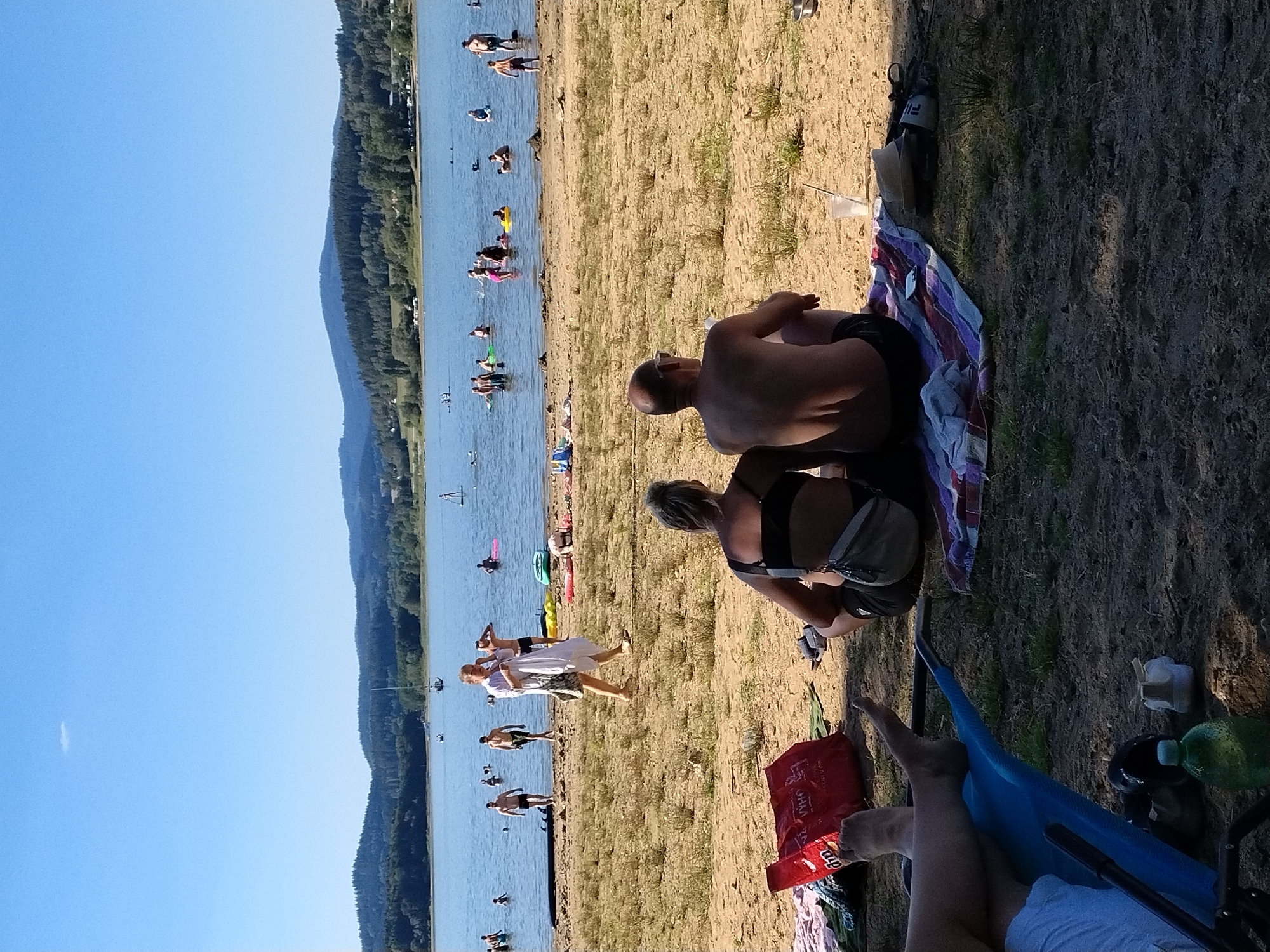 The image depicts a sunny day at a beach, likely in Radslav, Czechia, with people enjoying the water and the sand. In the foreground, a couple sits on a blanket facing the lake, with a red bag and other items scattered on the sand nearby. To the left, another person's legs are visible, resting on a blue chair.
The middle ground of the image features a sandy beach with scattered vegetation. Several individuals are visible walking on the beach or wading in the shallow water. A woman in a white cover-up walks away from the camera, carrying a white garment.
The background showcases a large body of water, a lake, filled with people swimming, floating on inflatable devices, and paddleboarding. Sailboats and distant trees and hills under a clear blue sky complete the scene, suggesting a popular recreational area. The overall atmosphere is one of a relaxed summer day.