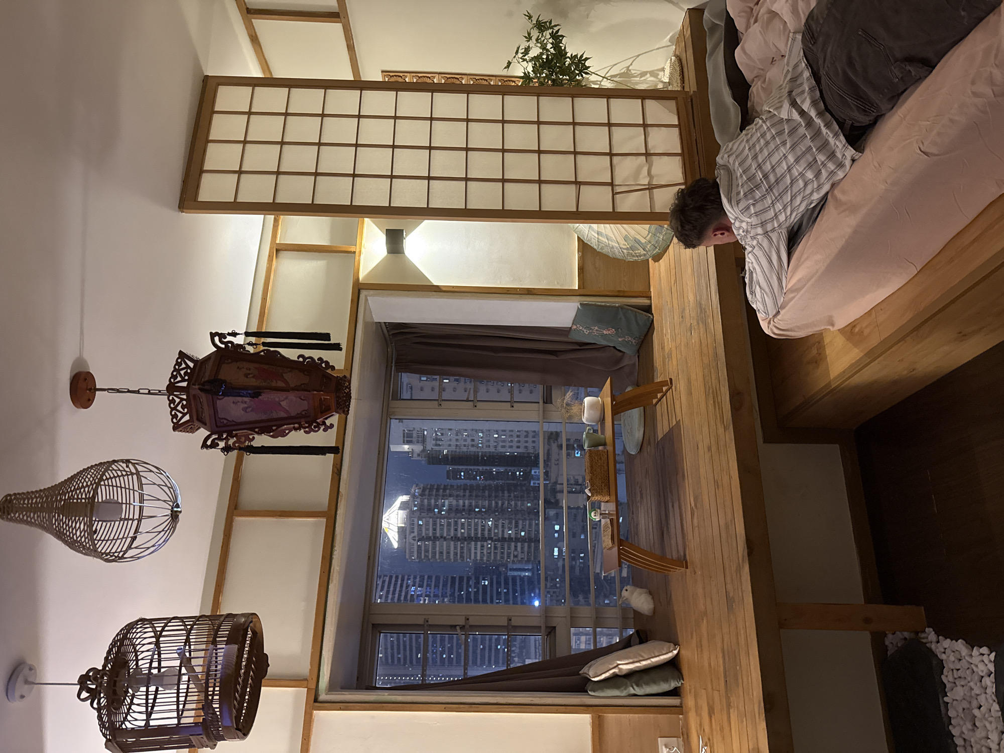 A person is lying on a bed in a room with a window overlooking the city of Yuzhong District, China. The room has Japanese-style interior elements, including a shoji screen and hanging wicker light fixtures. A multi-story building is visible through the window, with numerous windows illuminated at night. A narrow table with decorative objects is placed in front of the window.